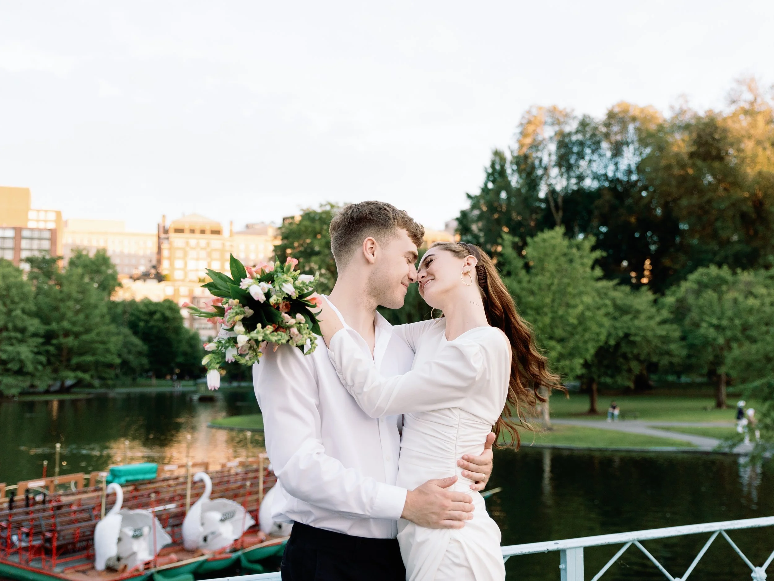 A couple in white outfits embracing on a bridge near a lake with swan-shaped pedal boats and lush green trees in the background.