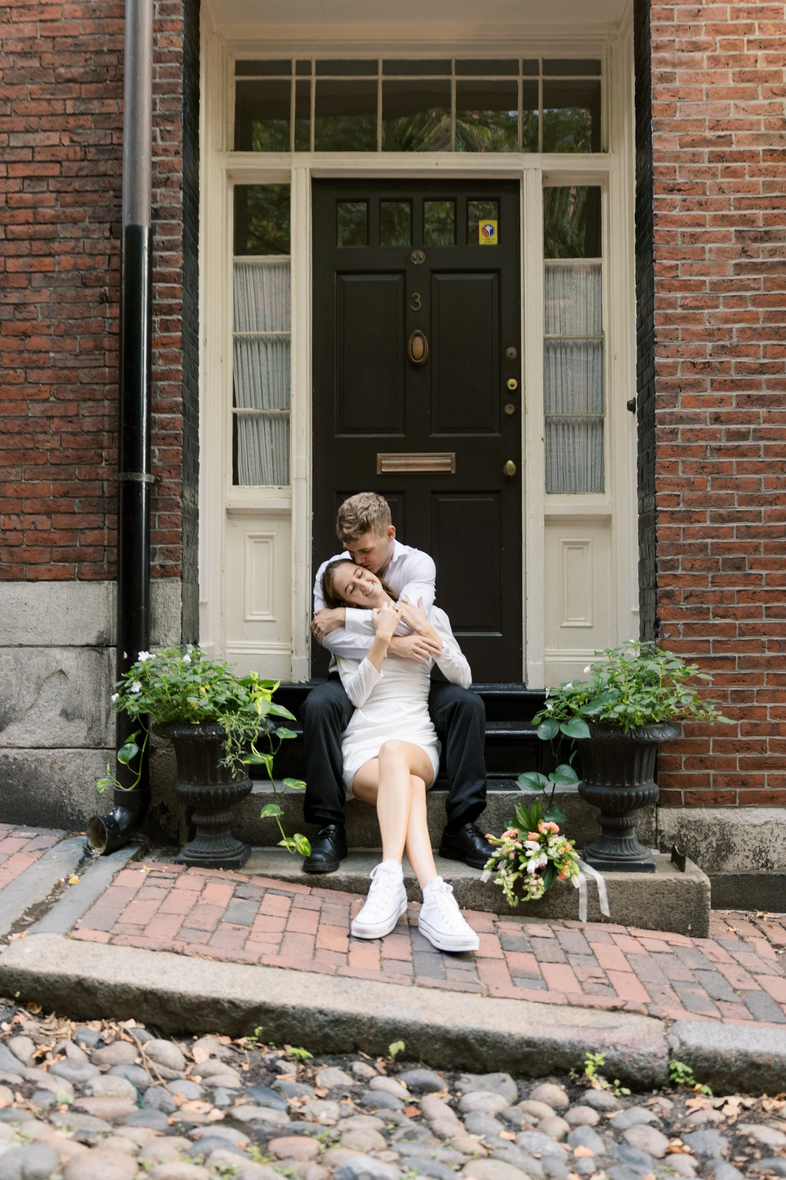 A young couple sitting on the front steps of a brick house, embracing lovingly; the woman is in a white dress and sneakers, and the man is in a white shirt and black pants. There are flower pots and a bouquet of flowers on the ground near them.
