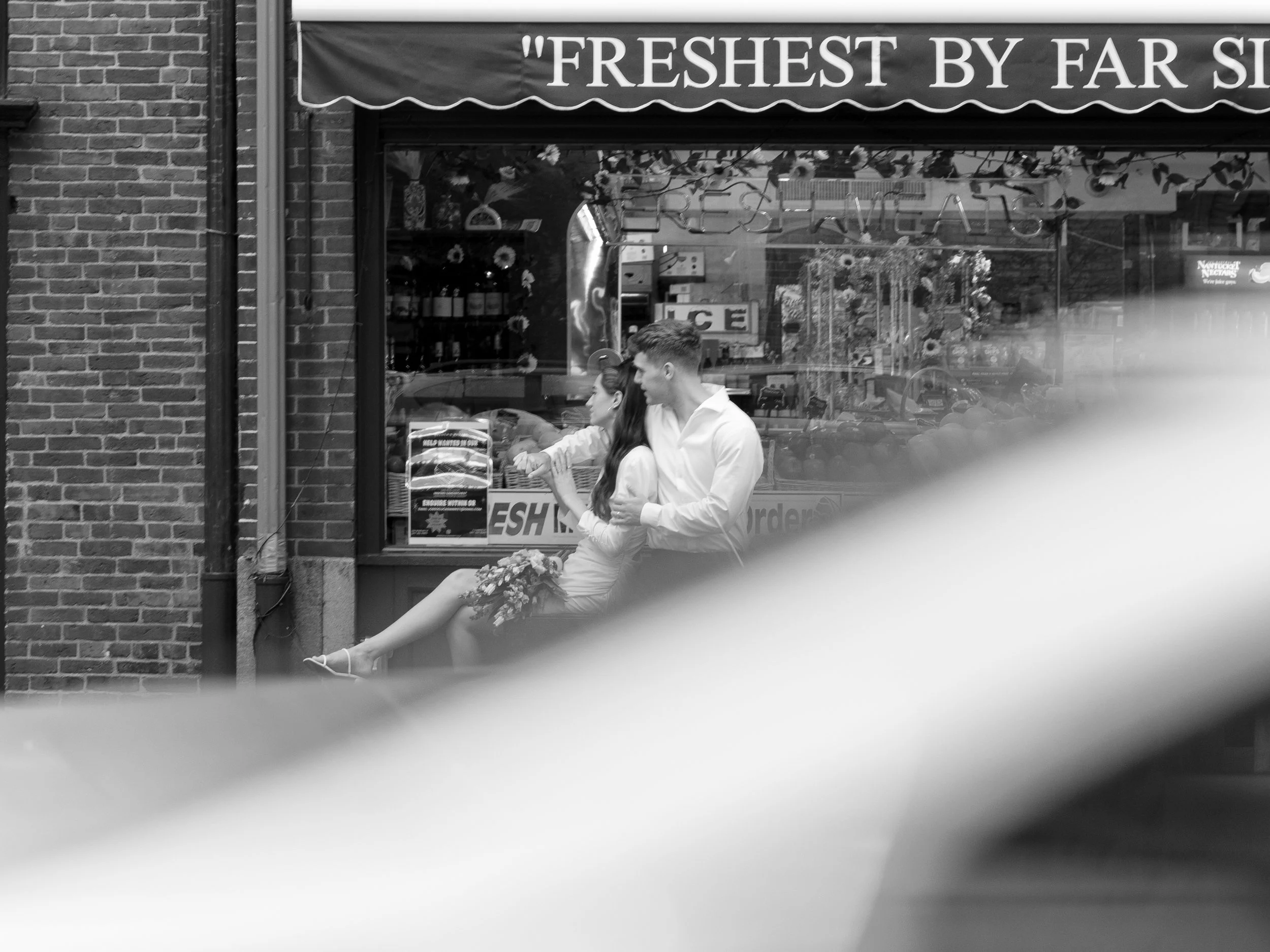 A black and white photo of a couple sitting outside a storefront with a sign that reads "FRESHEST BY FAR". The woman is holding a bouquet of flowers on her lap, and both are looking towards the street. The storefront has a display window with various