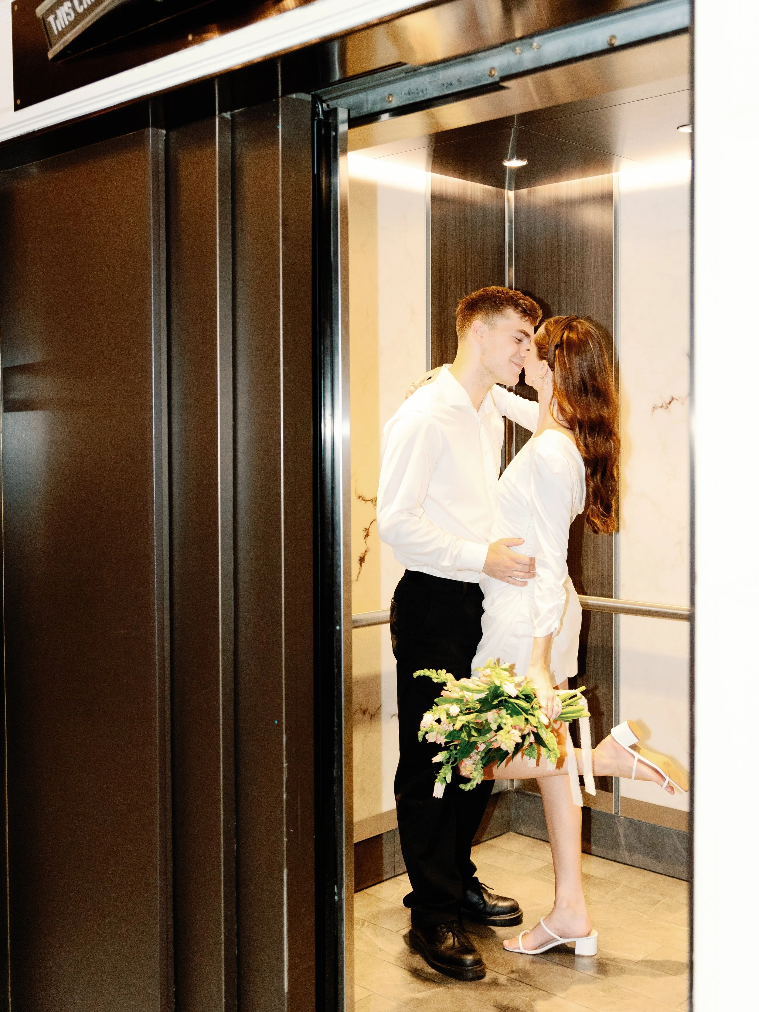 A young man and woman are in an elevator, sharing a romantic moment. The woman is holding a bouquet of flowers and her leg is lifted, showing her white high-heeled sandals.