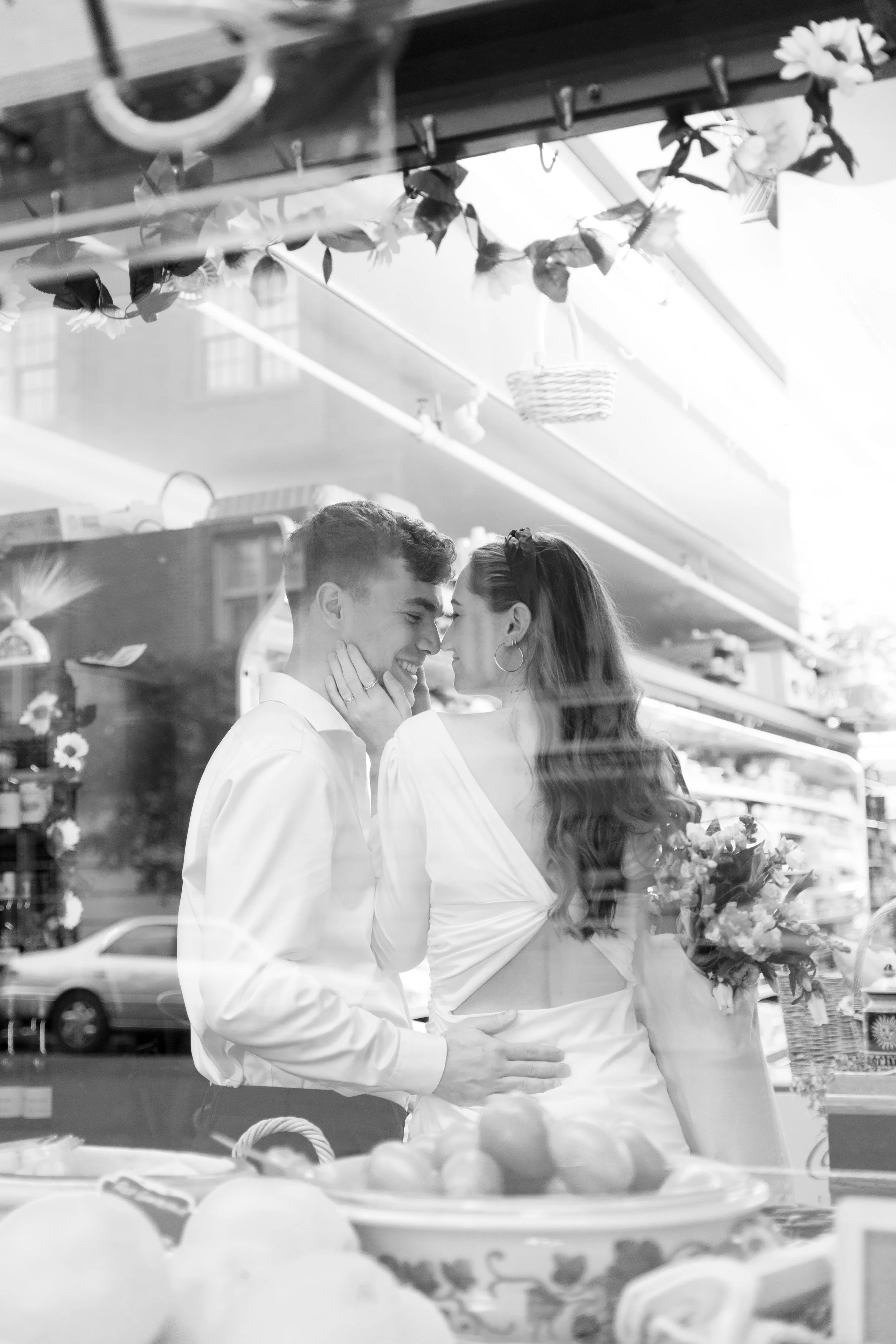 A black and white photo of a couple smiling and touching foreheads inside a store window display surrounded by fruits and flowers.