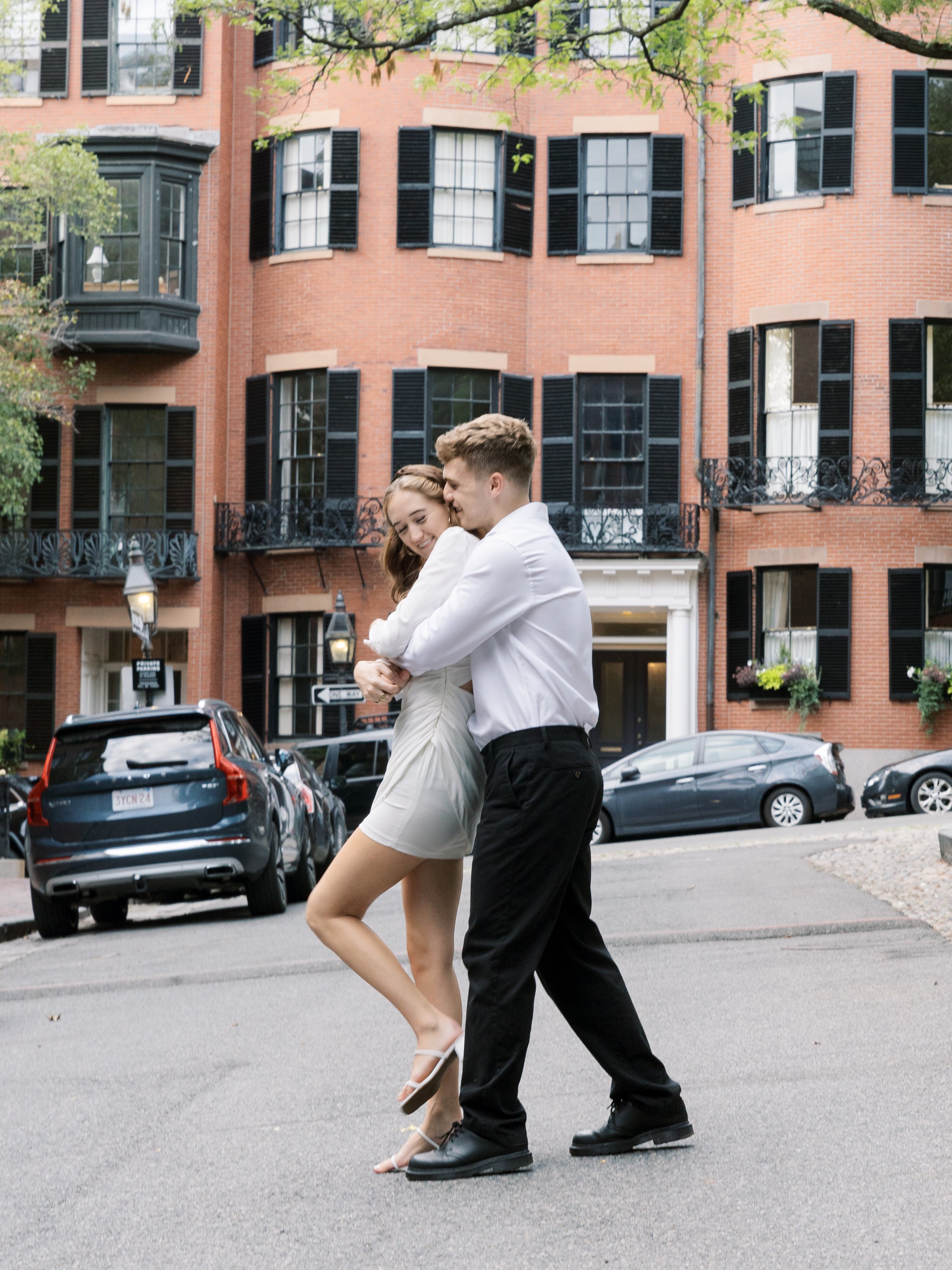 A young couple embracing and dancing in the street in front of a red brick apartment building with black shutters and balconies.