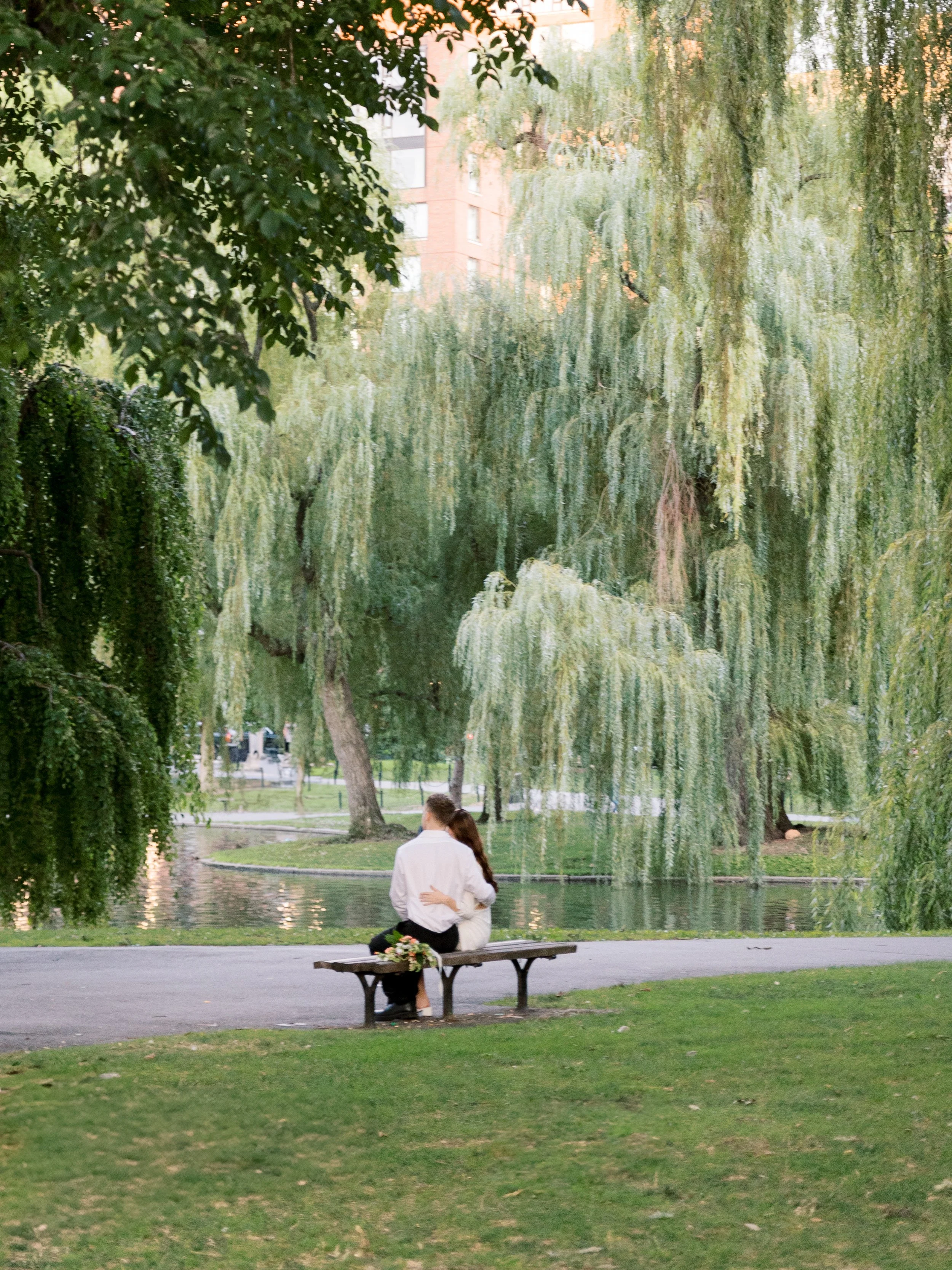A couple sits on a park bench near a pond with green trees surrounding them in a city park.