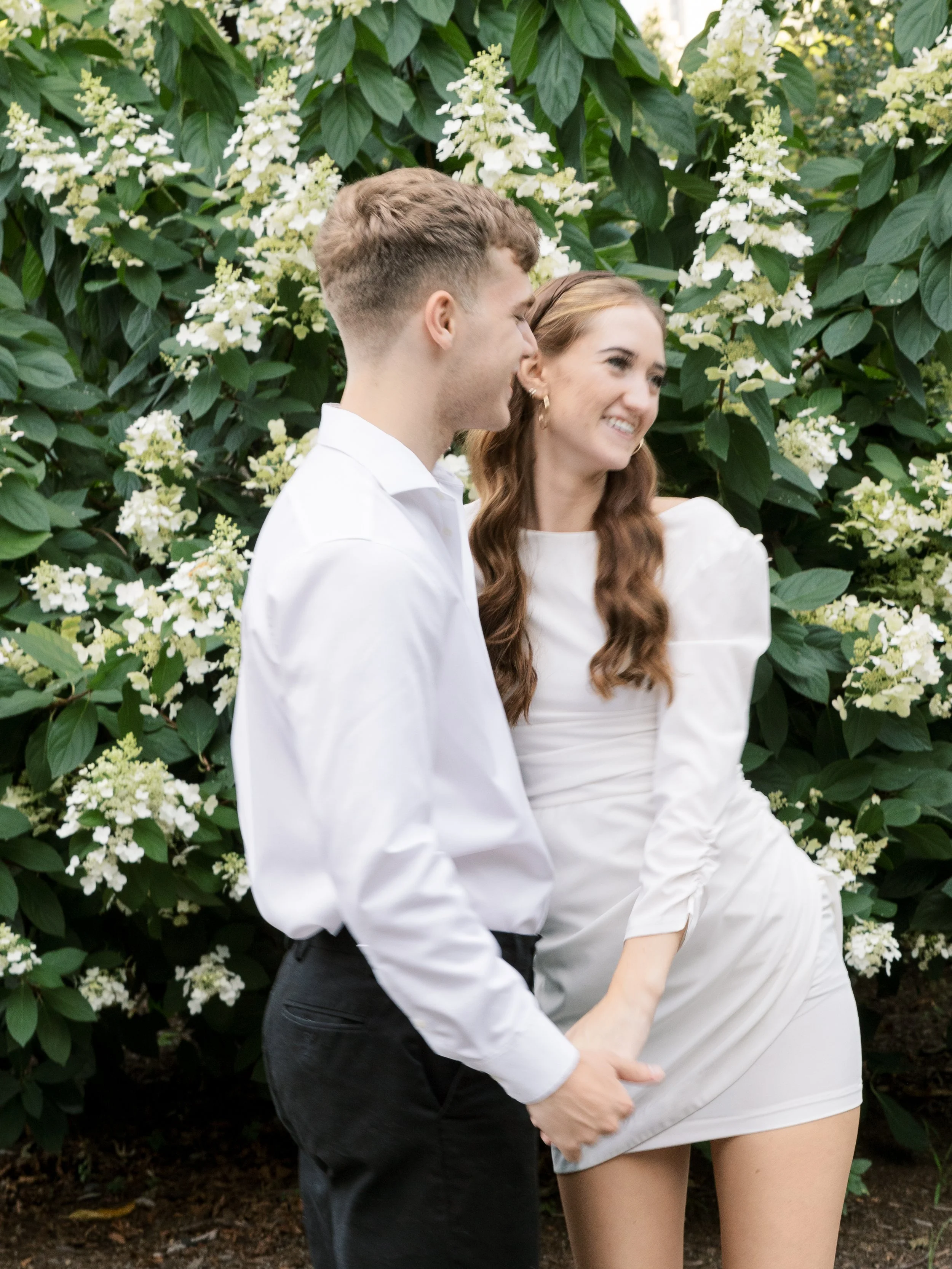 A young couple holding hands and smiling, standing in front of blooming white flowers and green foliage.