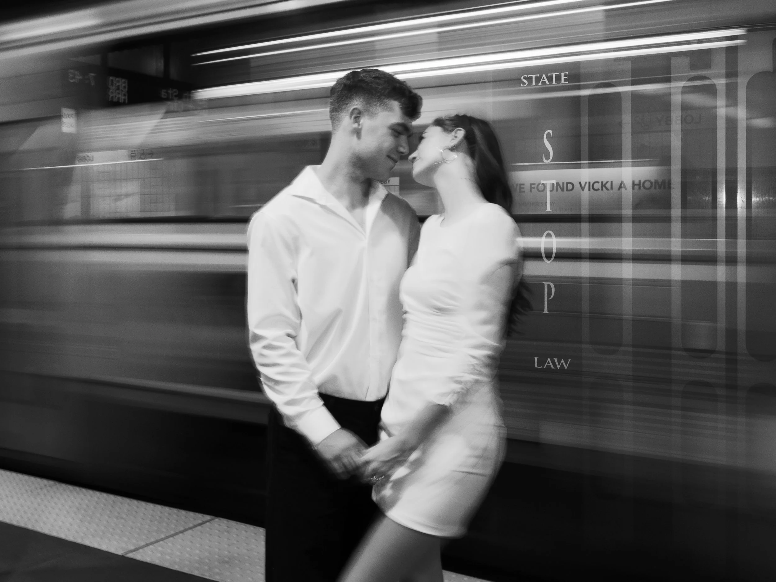 A black-and-white photo of a couple standing close together on a subway platform, about to kiss, with a moving train in the background.