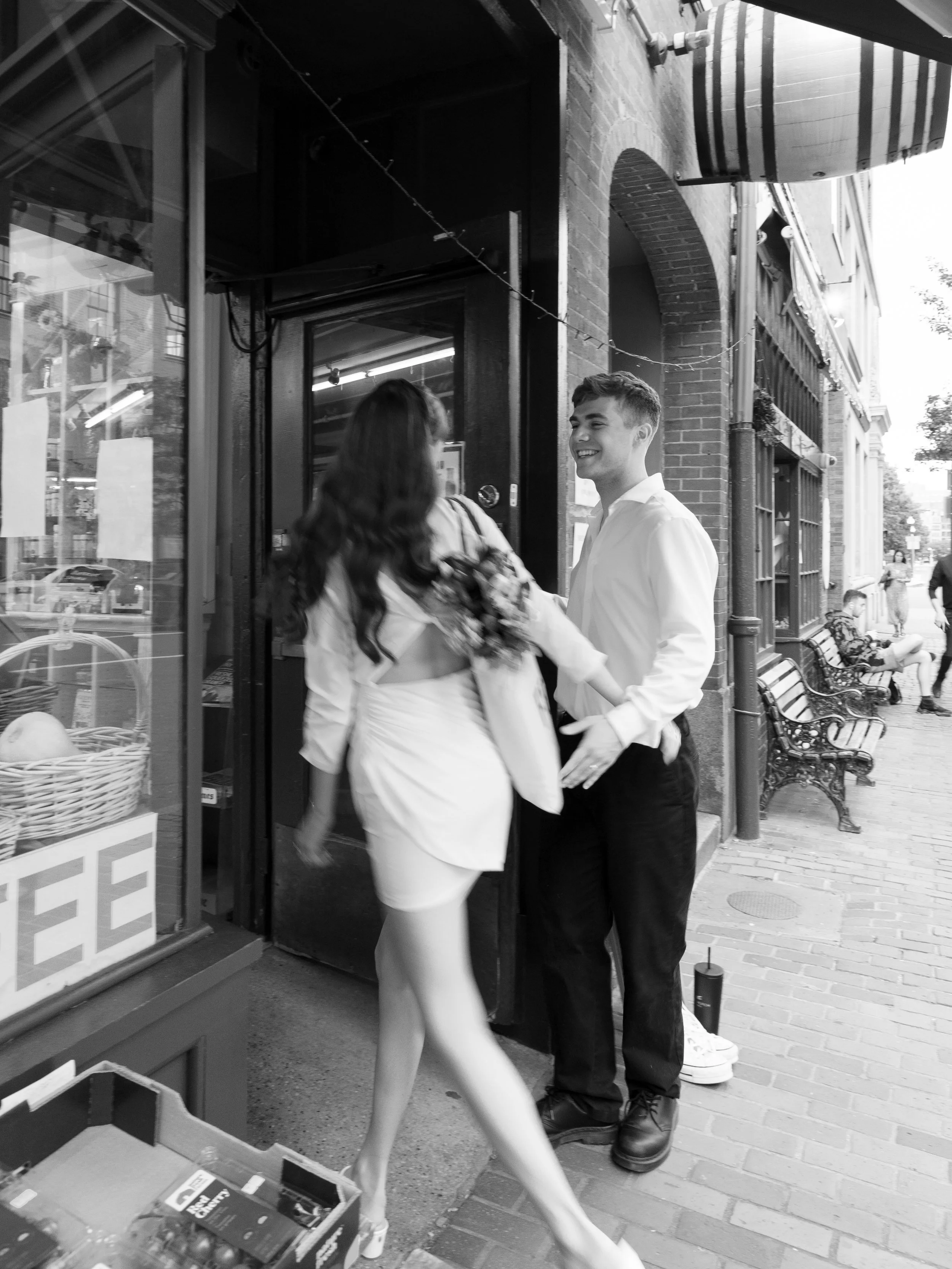 A young man and woman are smiling and talking outside a storefront, with the woman holding flowers and the man reaching out to her. The scene appears lively, with benches and pedestrians on the sidewalk.