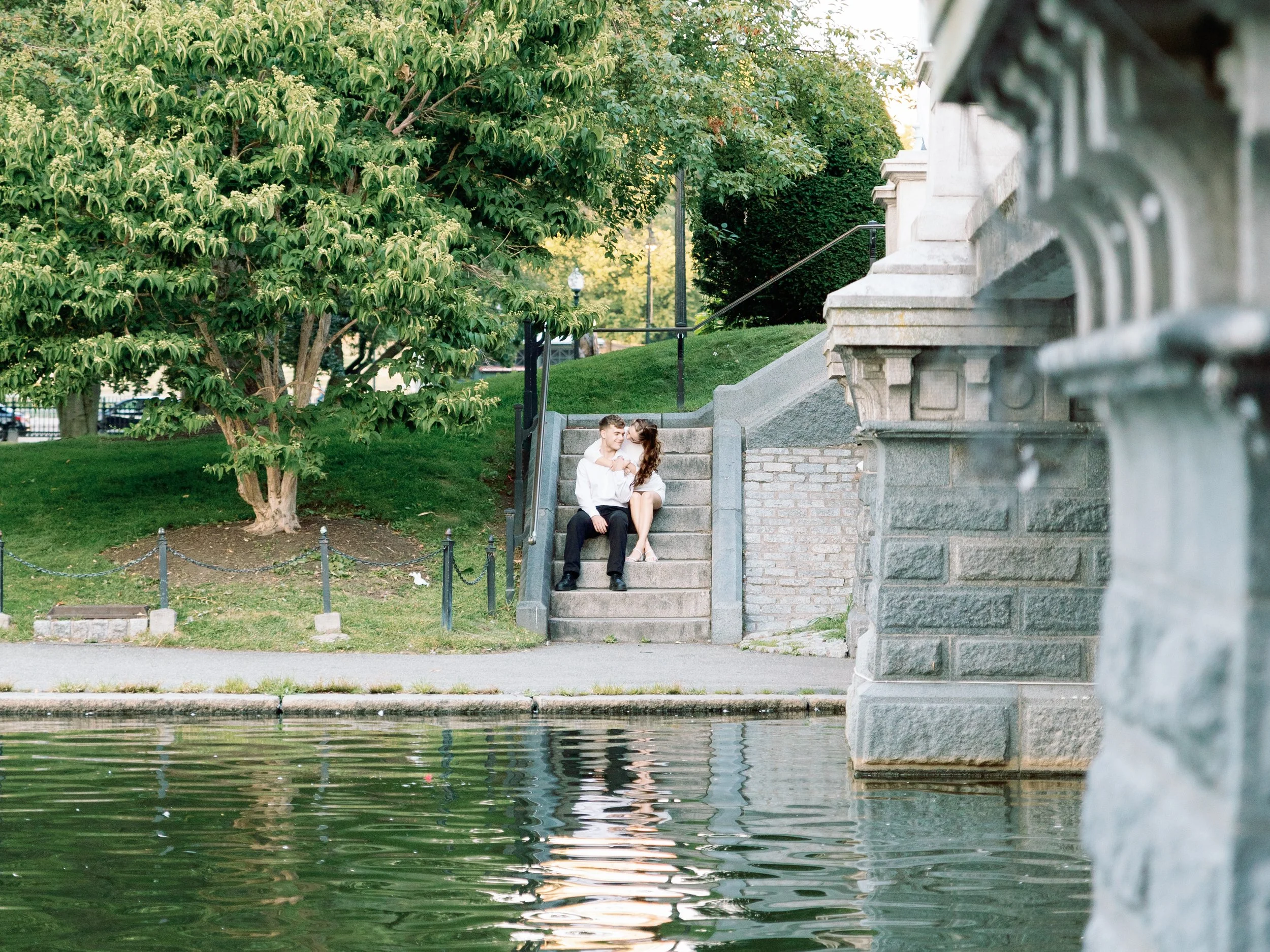 A couple sits on stone steps near a body of water, sharing a kiss, with greenery and a large tree in the background.