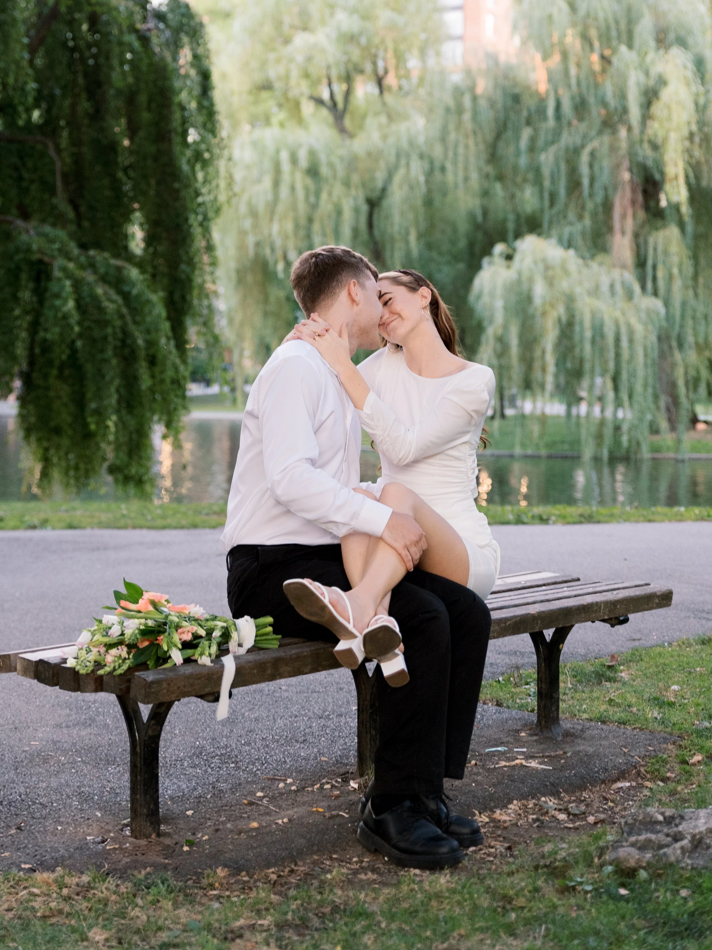 A couple sharing a kiss on a park bench, with a bouquet of flowers nearby, during daylight with greenery and trees in the background.