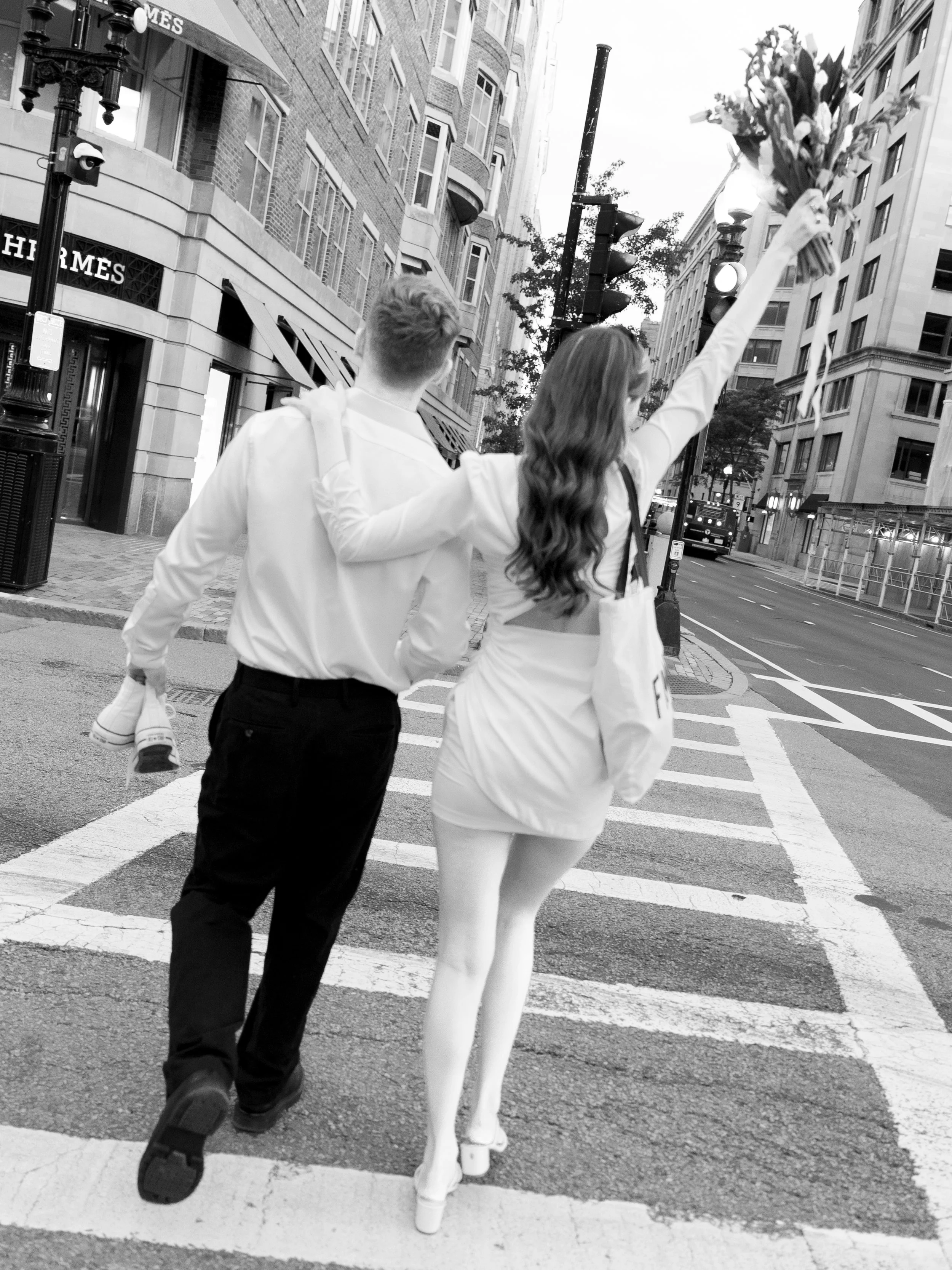 A young couple crossing a city street at dusk; the woman with long hair in a skirt and heels holds a bouquet of flowers.