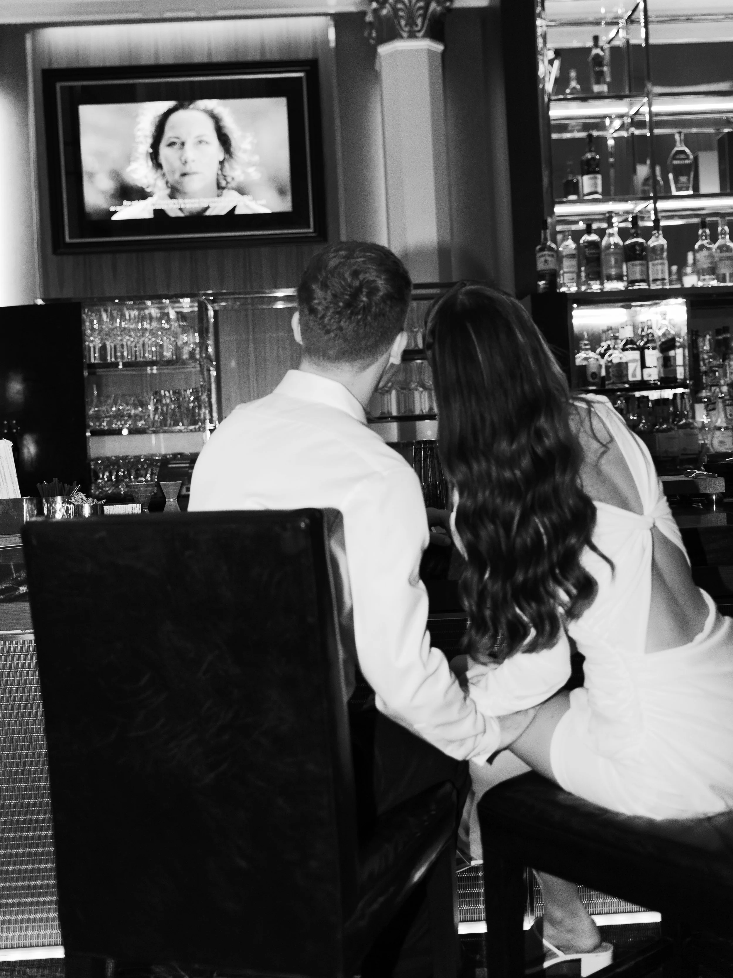 A man and woman sitting closely together at a bar, watching a TV screen in a black-and-white photo.