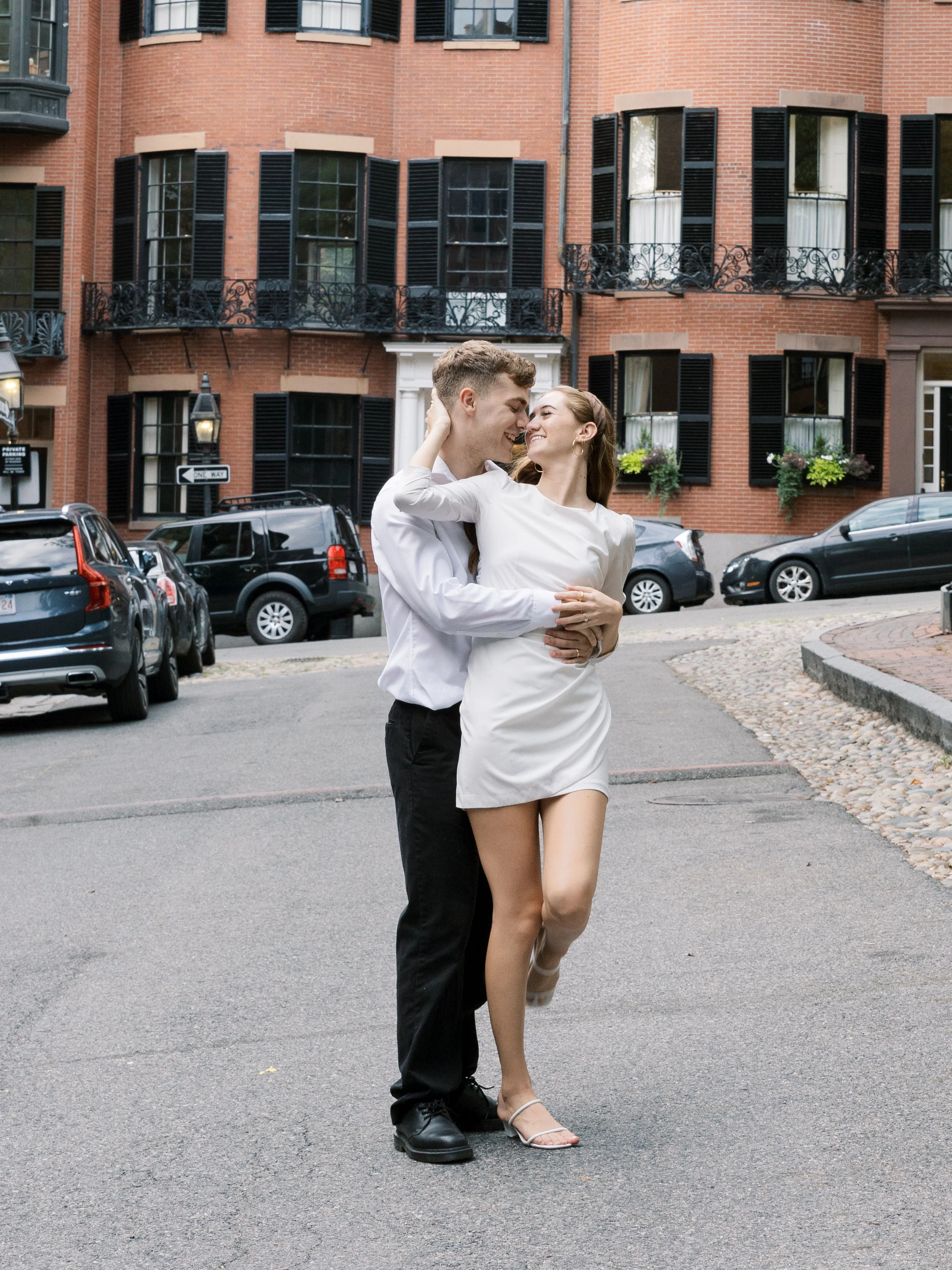 A young couple dancing joyfully outdoors on a city street with brick buildings and cars parked along the curb in the background.