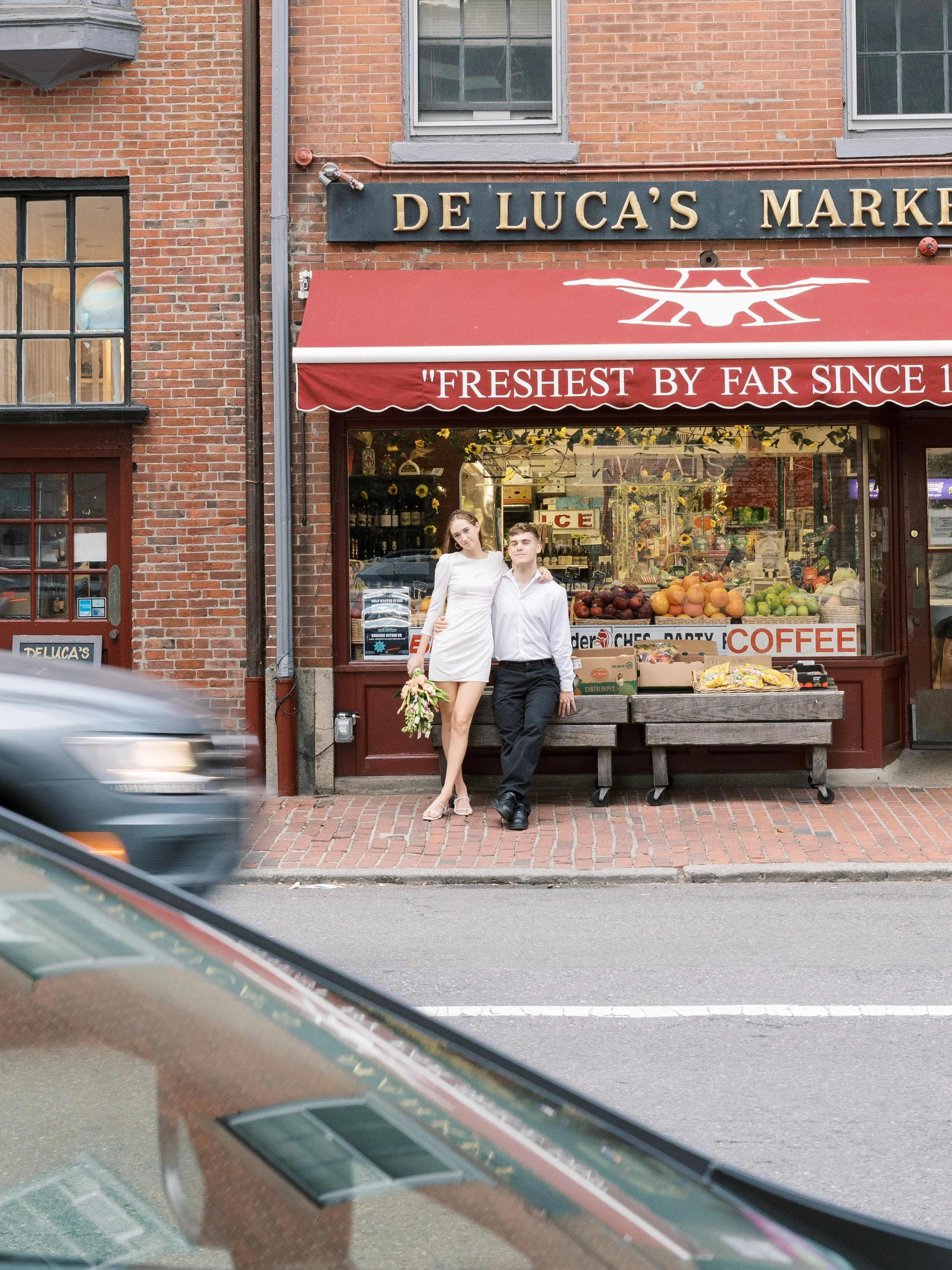 A young couple standing outside a historic brick-market store, with the woman holding a bouquet of flowers, and the store's display window filled with produce and signs.