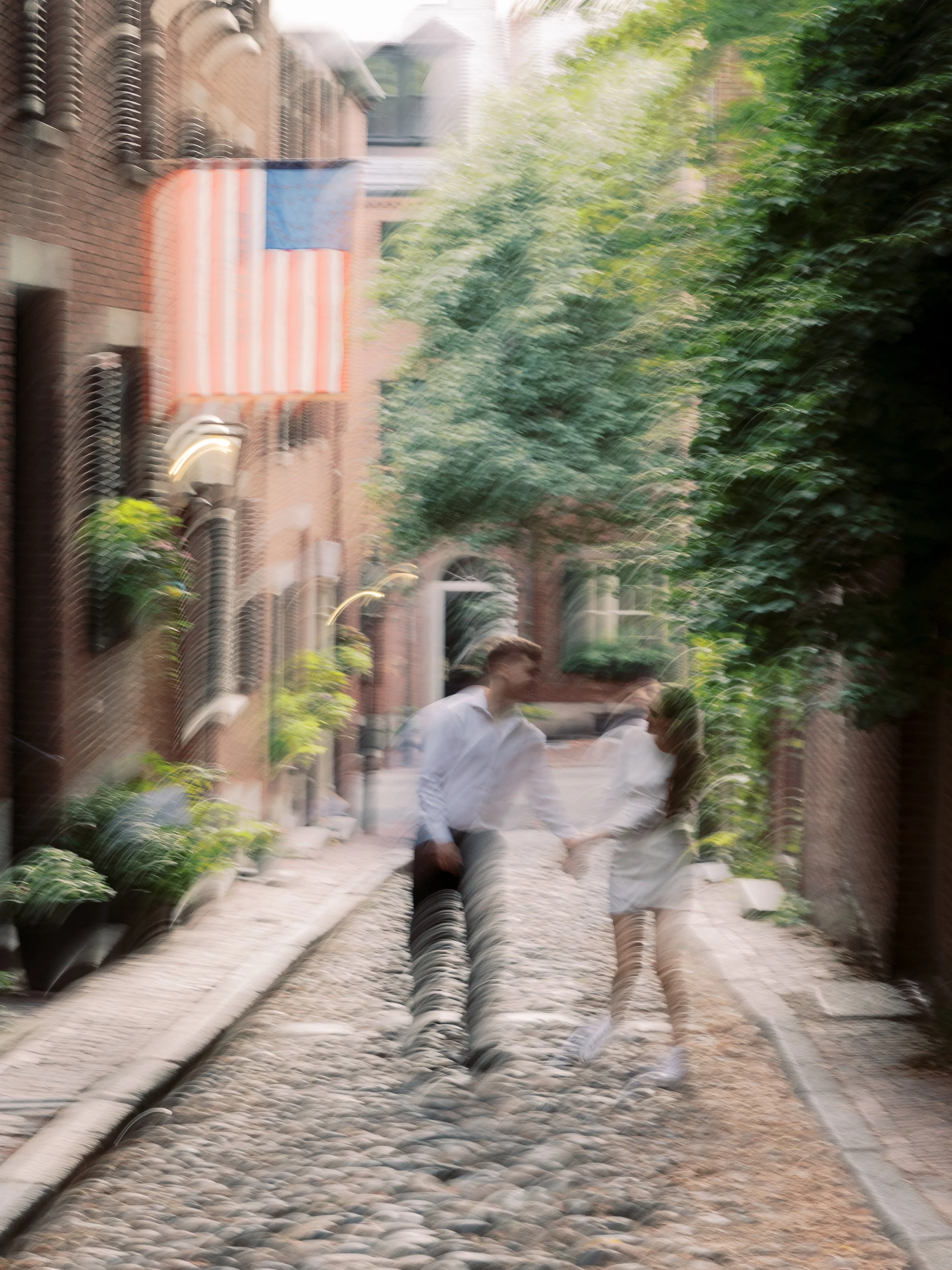 A blurry image of a couple holding hands and walking down a cobblestone street lined with brick buildings and green trees, with an American flag hanging on the building.