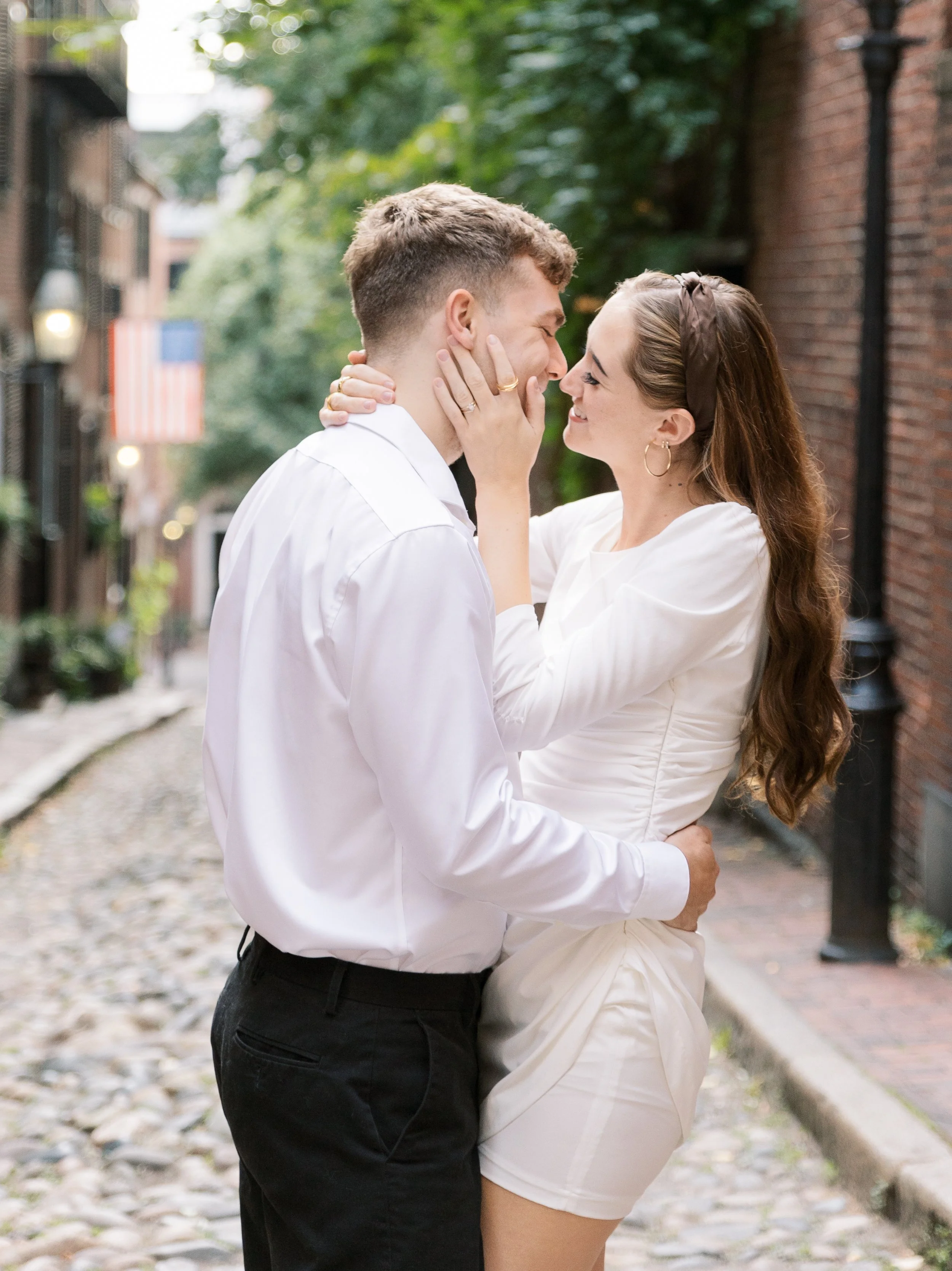 A couple standing close together outdoors on a cobblestone street, smiling and touching noses, with a background of buildings, trees, and an American flag.