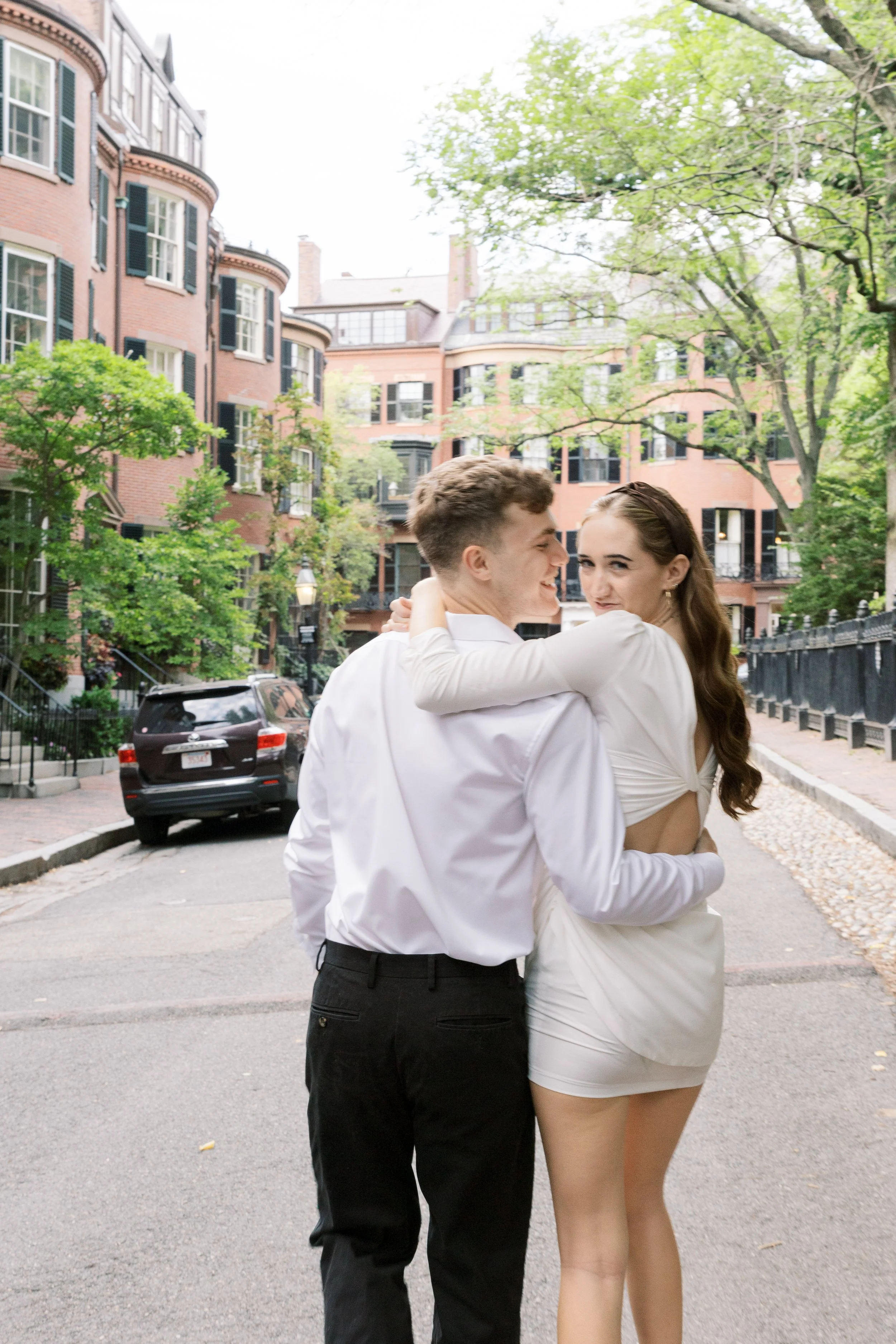 A couple hugging on a city street surrounded by brick buildings and trees.