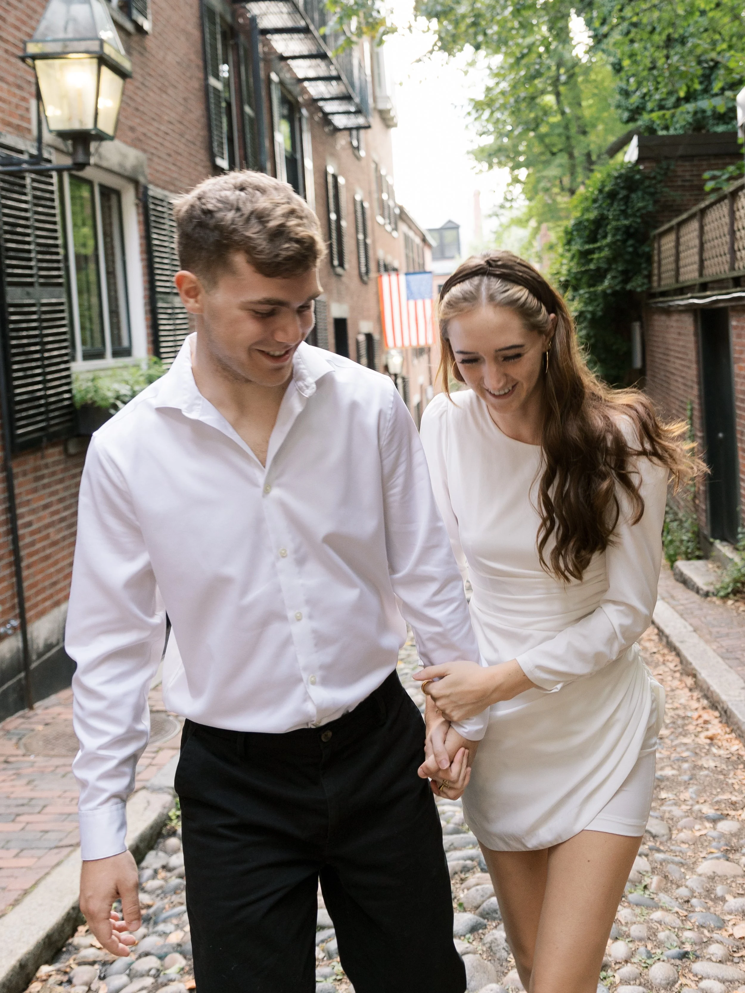 Young couple walking arm-in-arm on a cobblestone street in a residential neighborhood, smiling and enjoying each other's company.