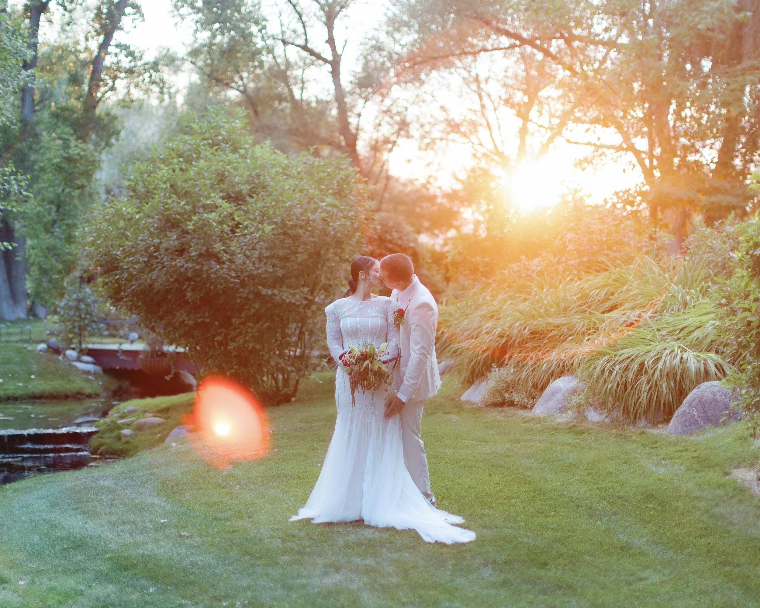 A bride and groom sharing a kiss in a garden during sunset, with lush greenery and rocks in the background.