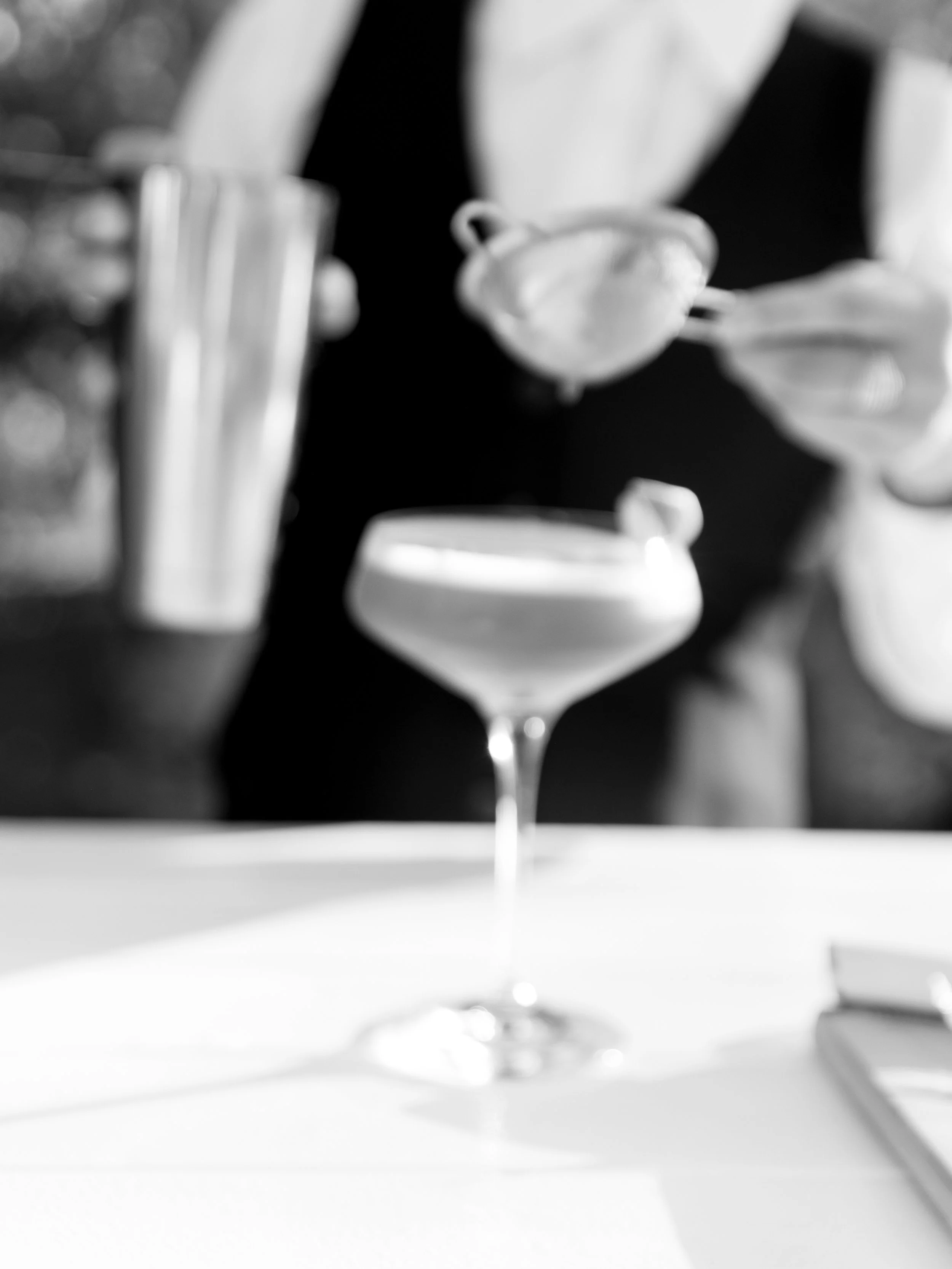 Black and white photo of a martini glass with a cocktail, blurred background with person holding a cocktail strainer.
