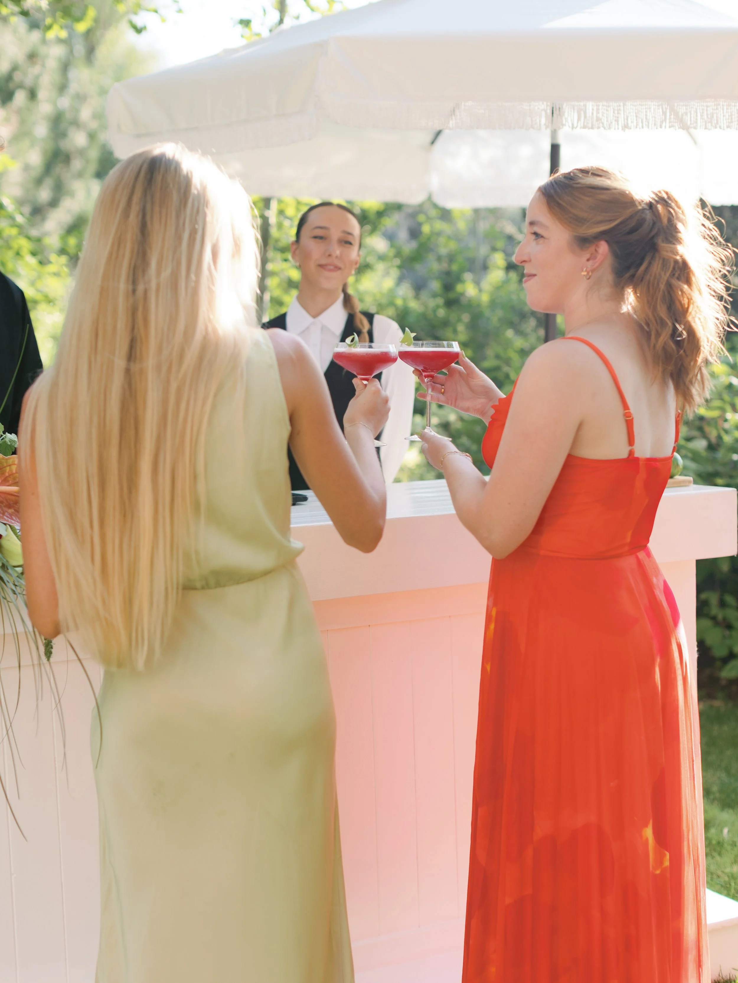 Two women in colorful dresses clink glasses of red cocktail at an outdoor event next to a white bar under a large white umbrella.