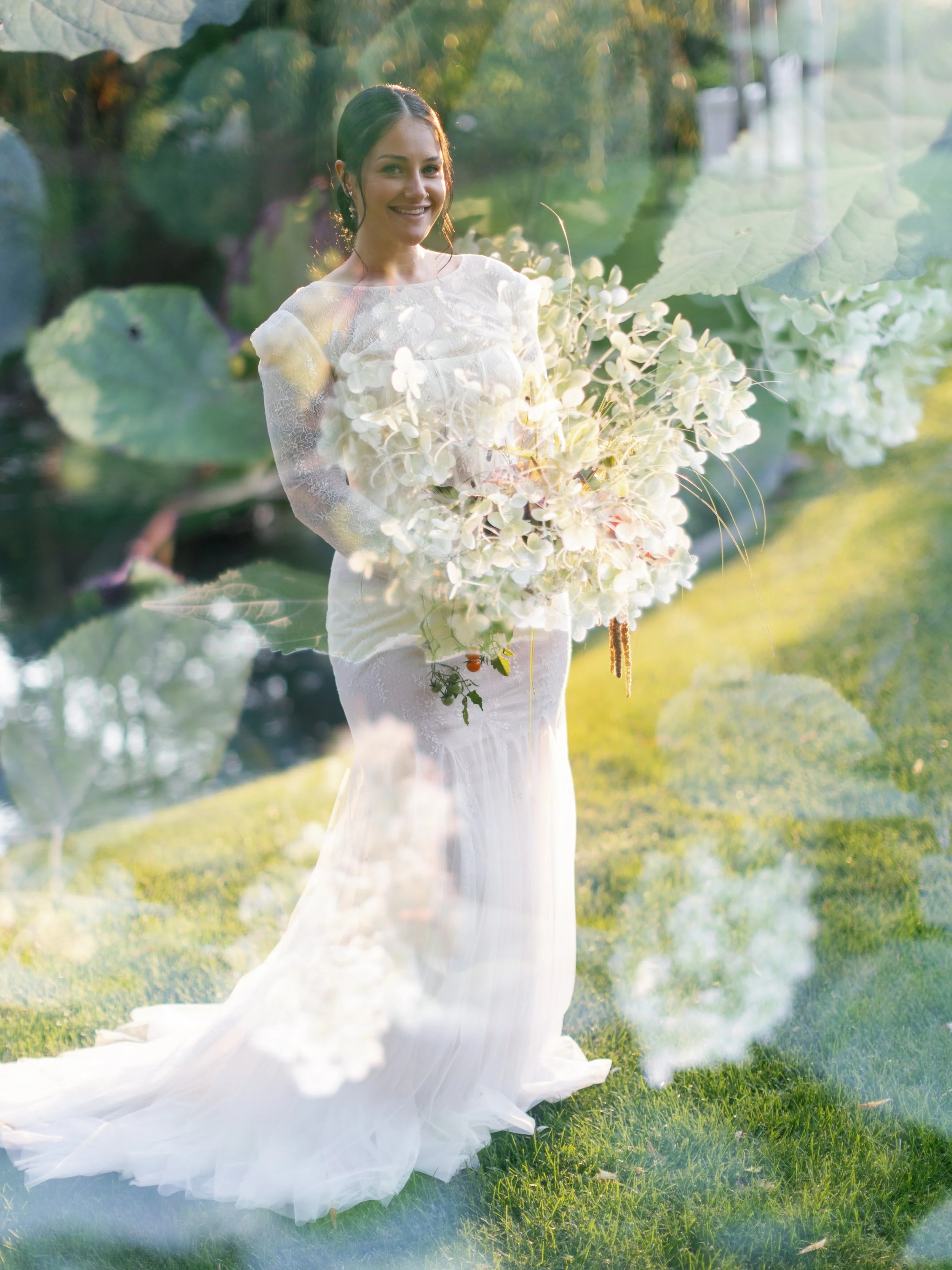A woman in a white wedding dress holding a large bouquet of white flowers, photographed outdoors in sunlight, with a double exposure effect of greenery and flowers.