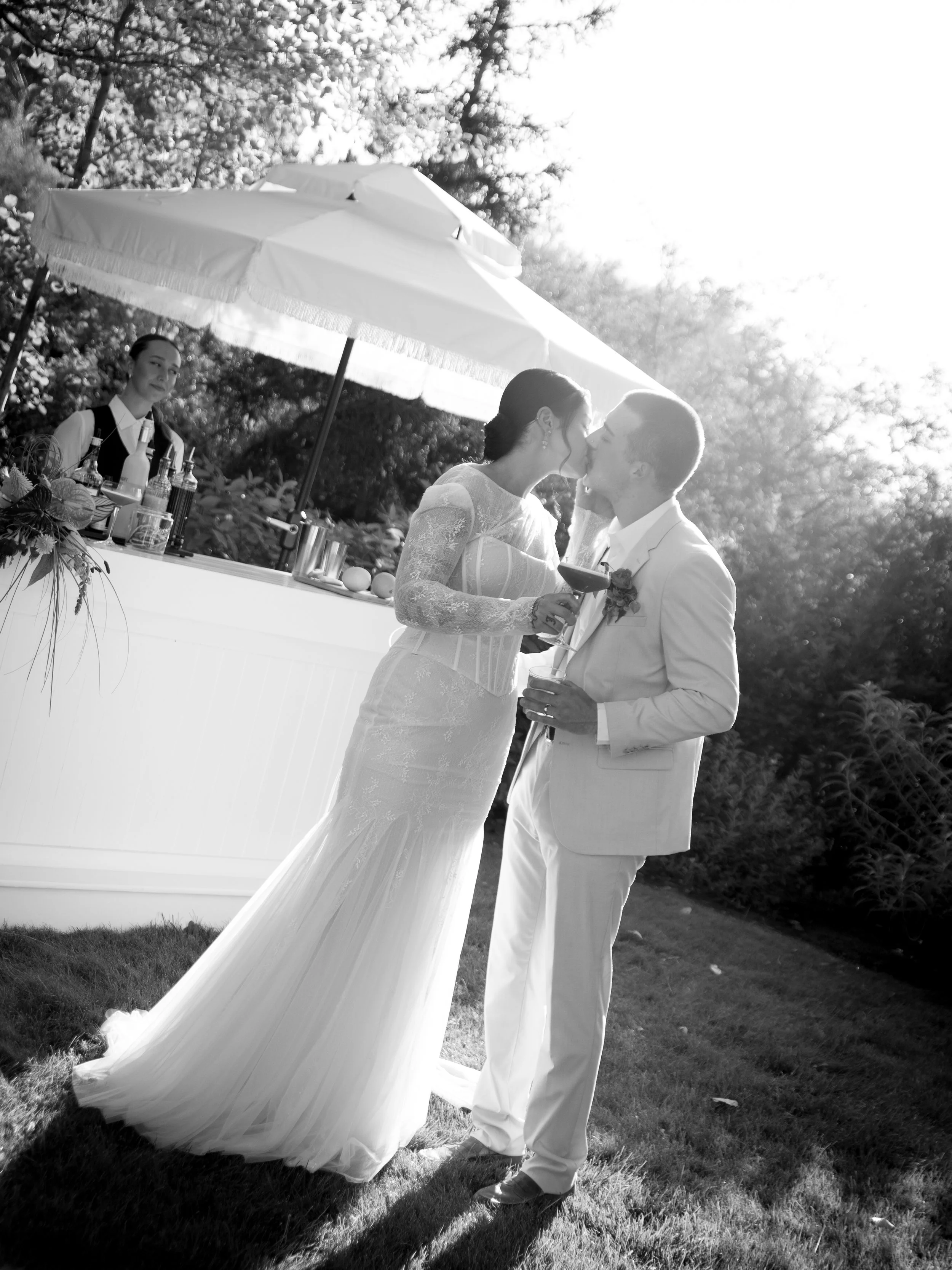 A black-and-white photo of a bride and groom sharing a kiss during their outdoor wedding reception, with a bar and a bartender in the background under a large umbrella.