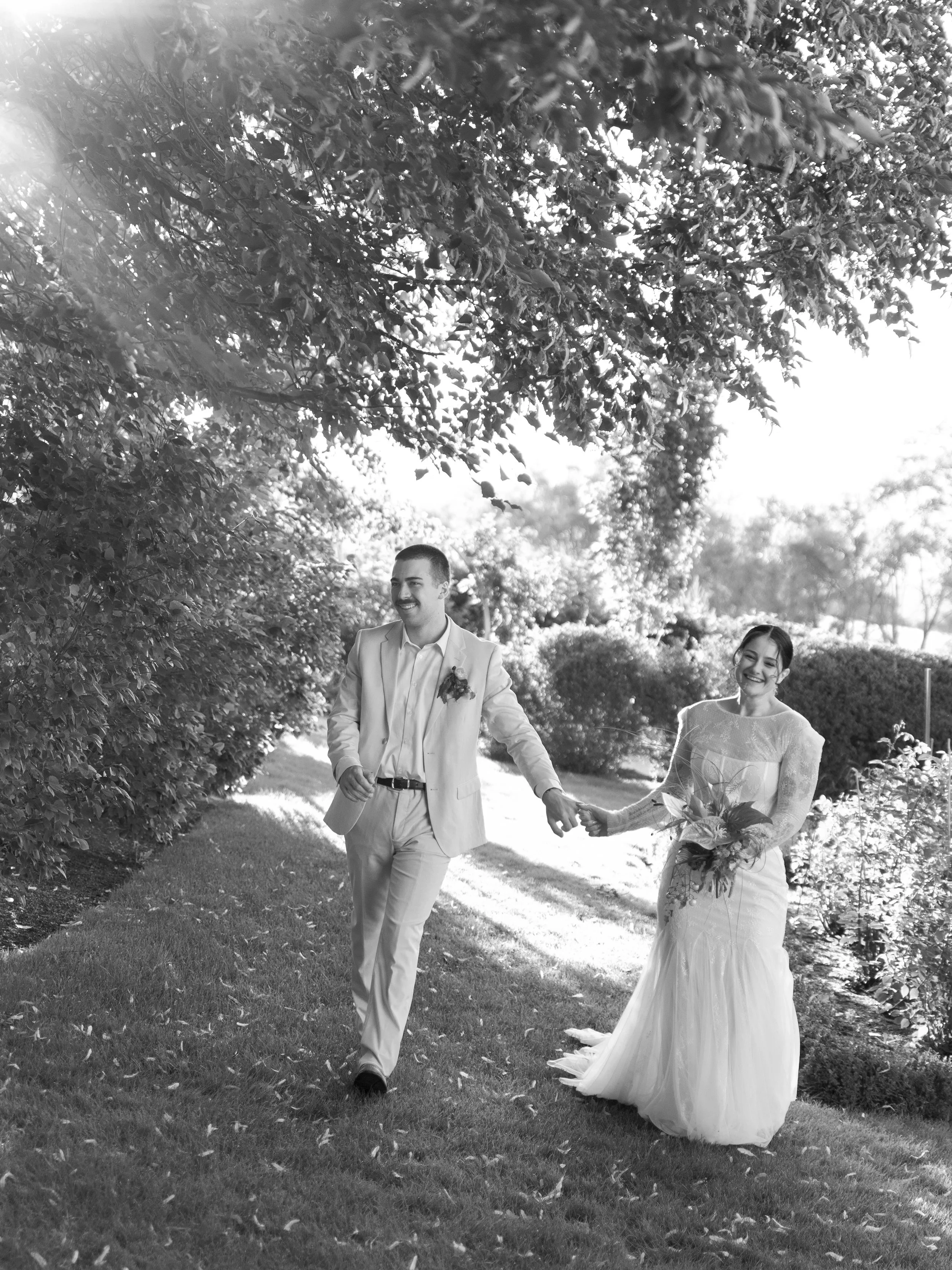 Black and white photograph of a newlywed couple walking hand in hand through a garden, smiling, with lush trees and bushes around them.