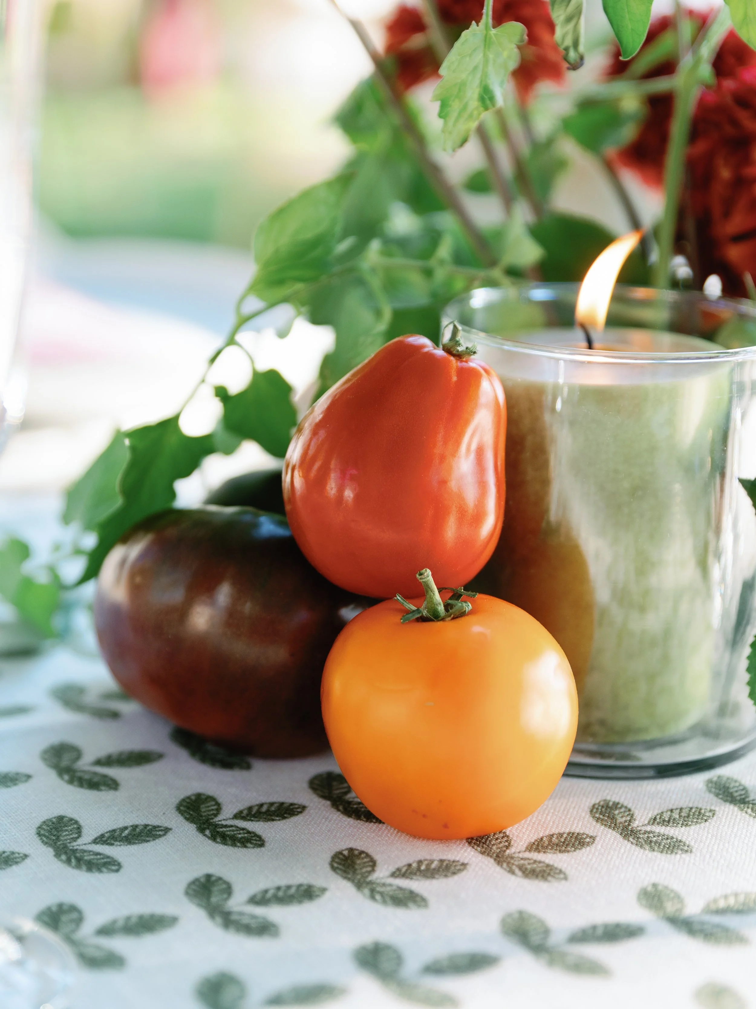 Close-up of heirloom tomatoes in various colors and a green candle in a glass holder on a patterned tablecloth, with green leaves and blurred outdoor background.