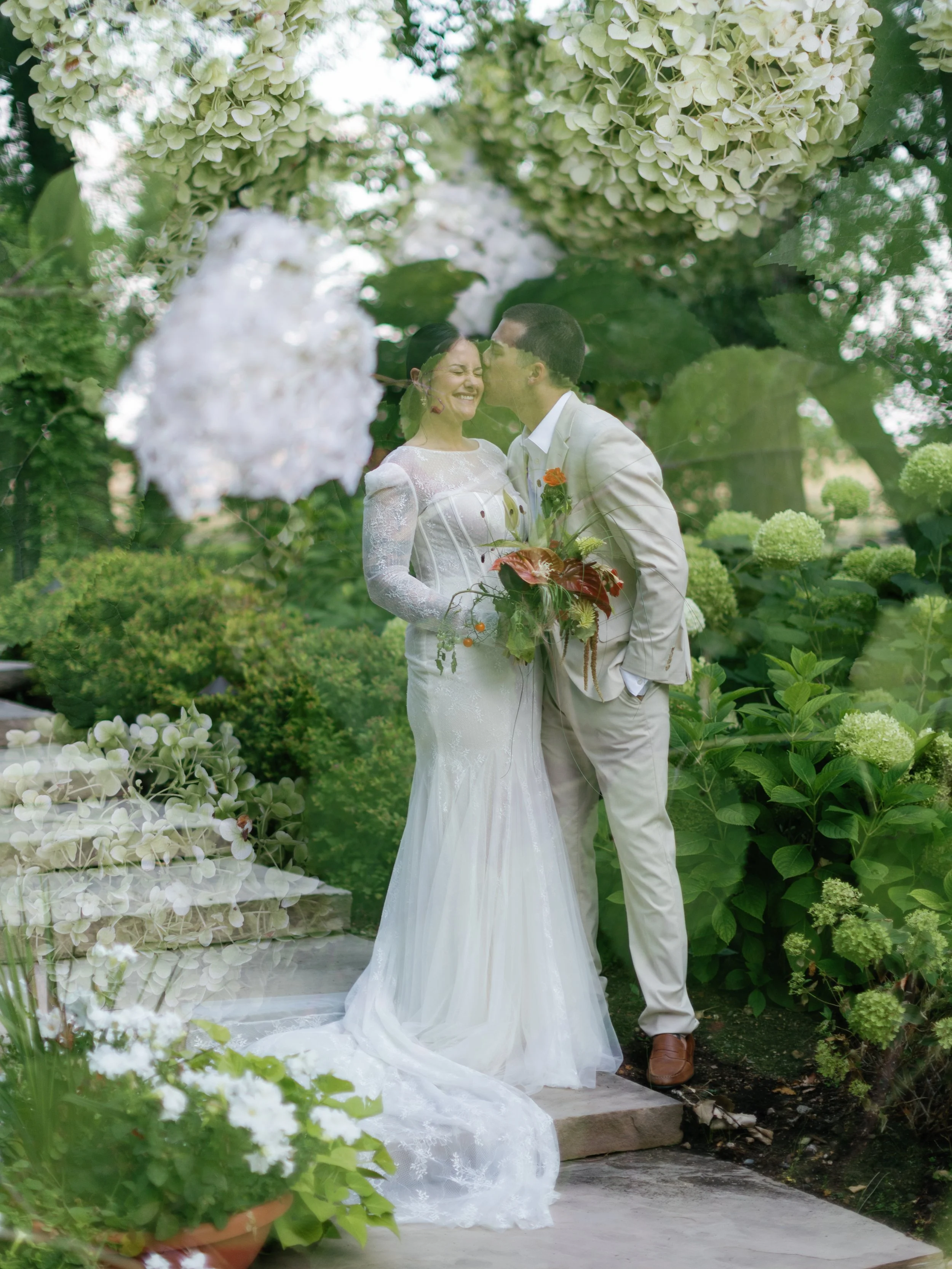 A bride and groom sharing a joyful moment in a lush garden, with the groom leaning in to kiss the bride on the forehead, surrounded by green foliage and white flowers.
