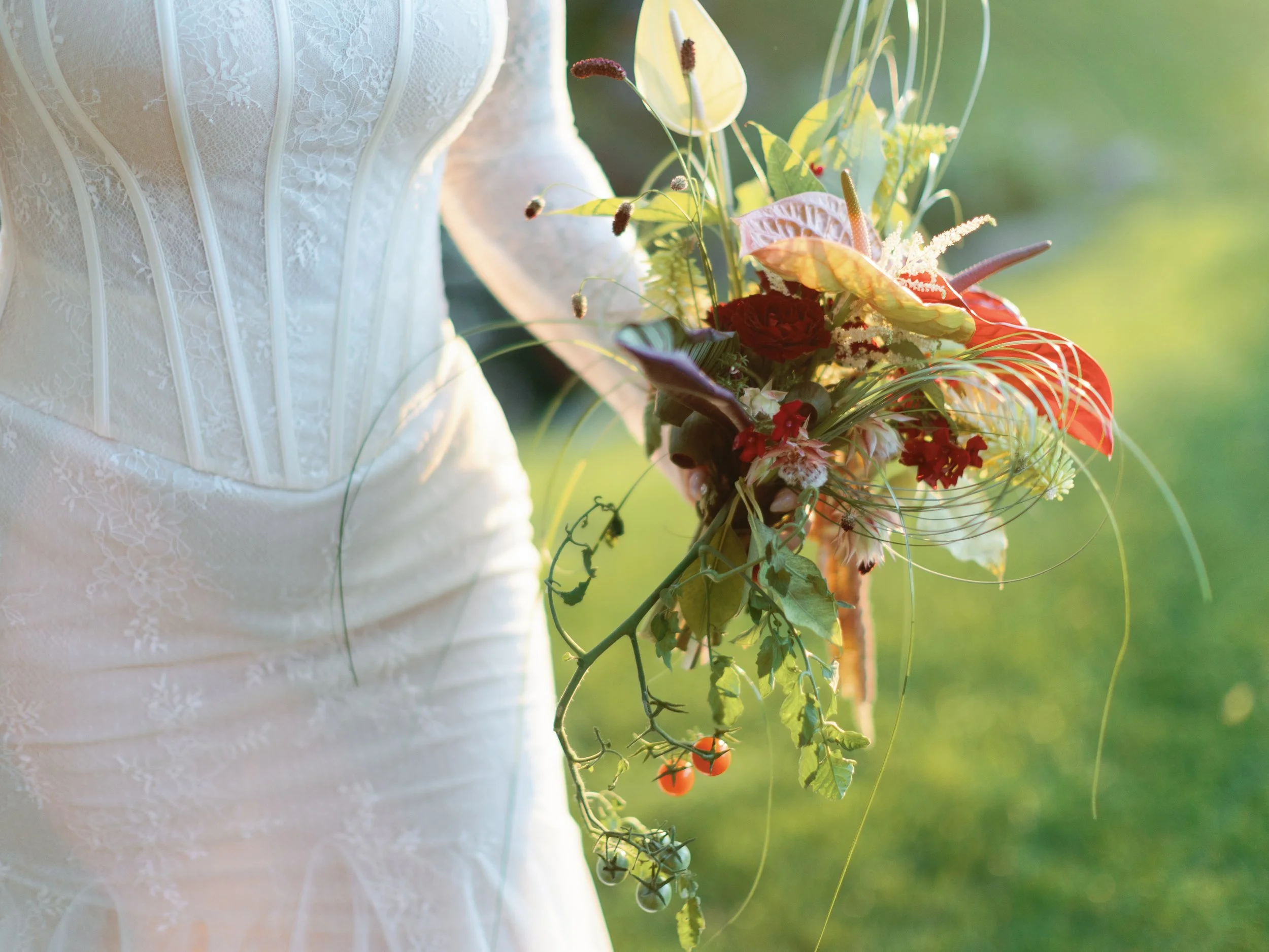 Close-up of a person in a white lace wedding dress holding a colorful bouquet of flowers outdoors, with greenery in the background.