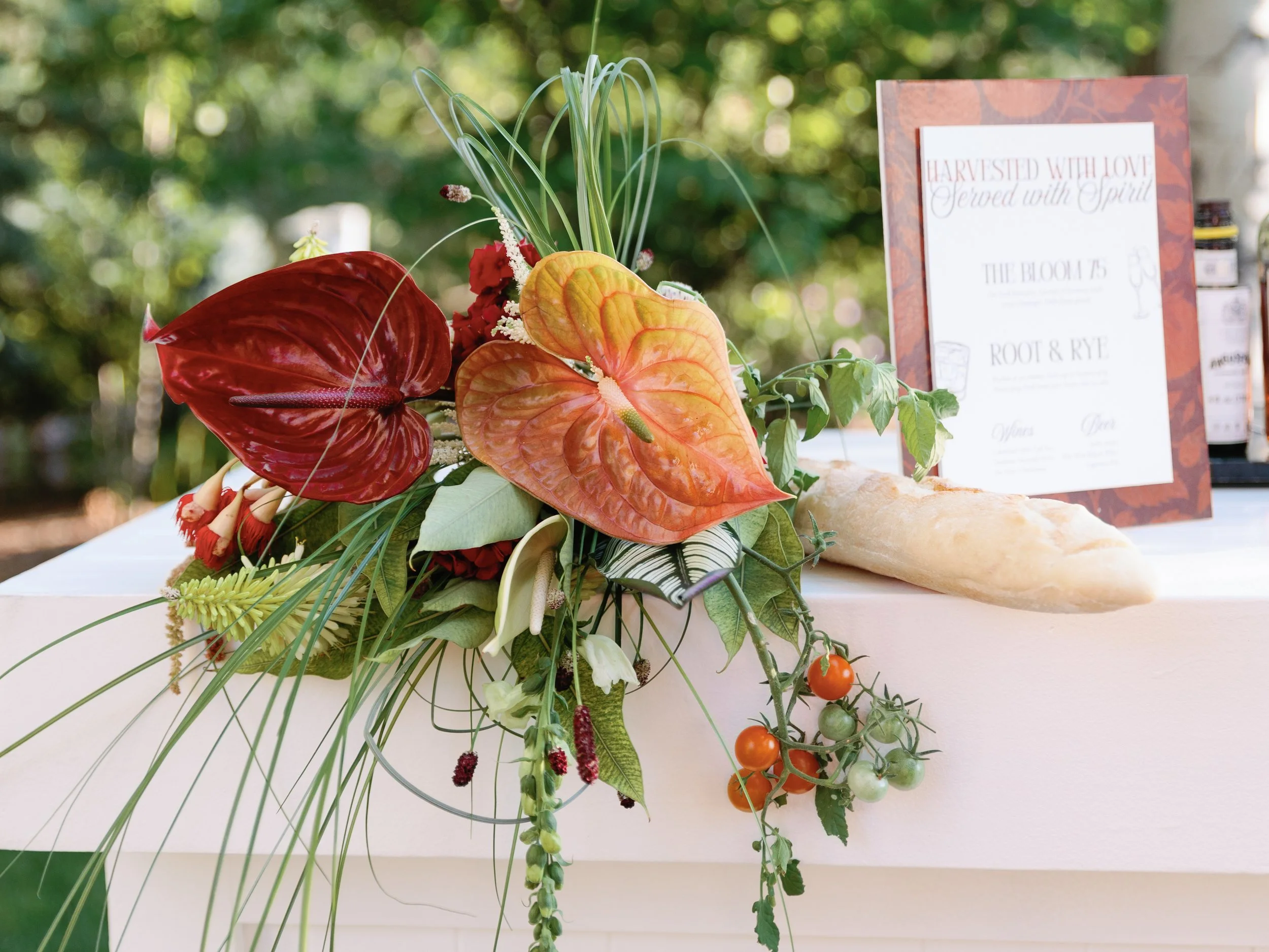 A floral arrangement with colorful anthuriums, greenery, and small tomatoes, placed on a white table with a bread loaf and a framed menu in the background at an outdoor event.