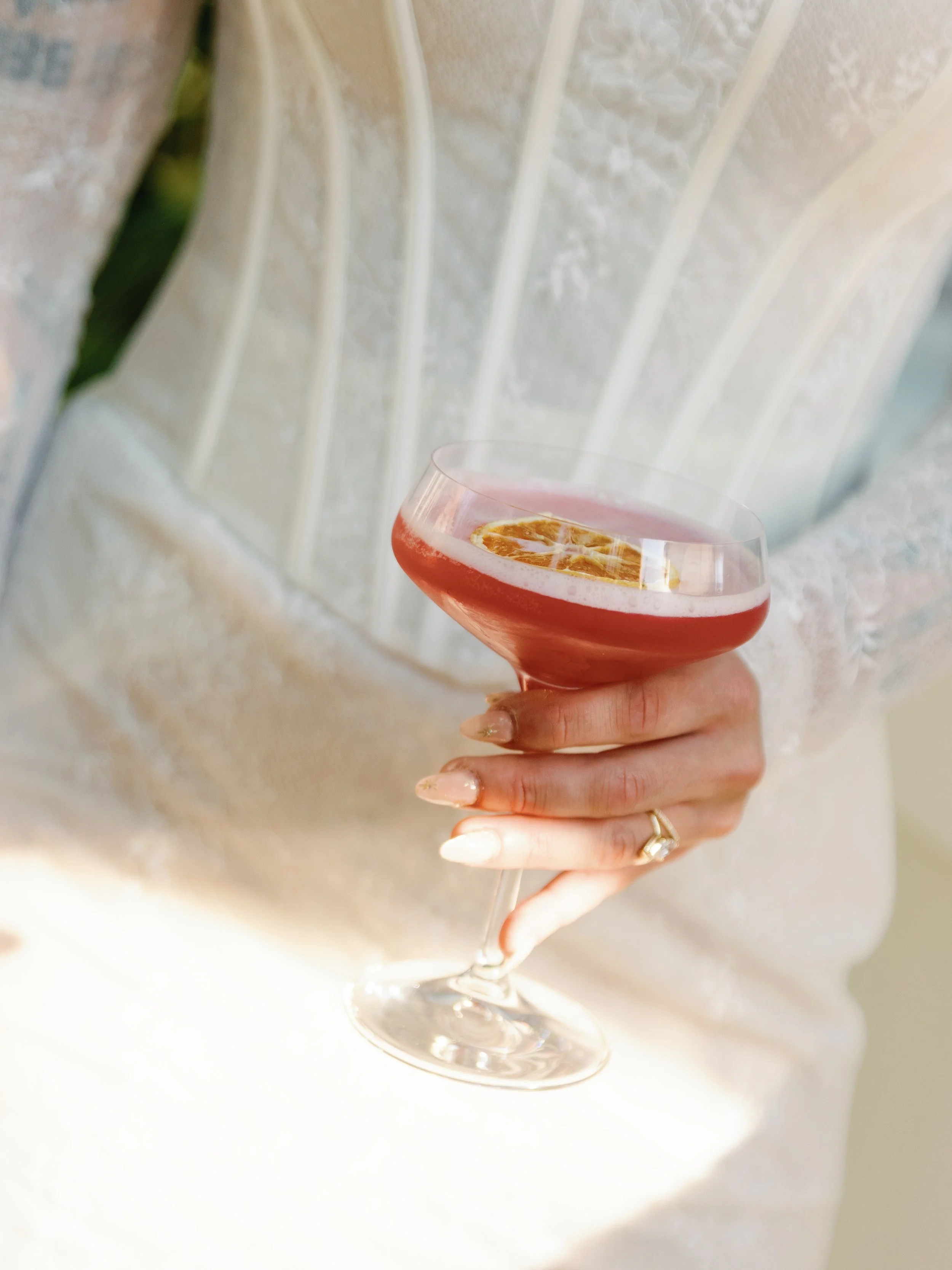 A person in a wedding dress holding a glass of pink cocktail with a lemon slice garnish.