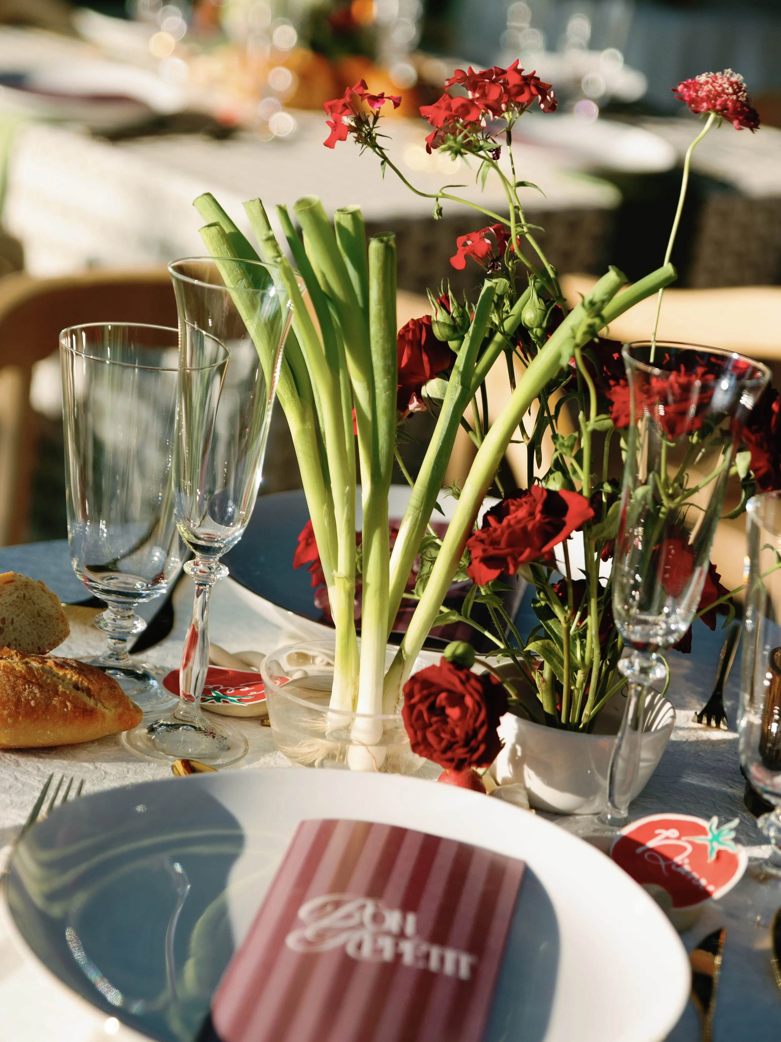 Table setting with a centerpiece of red and pink flowers, wine glasses, bread, and a striped napkin on a white plate.