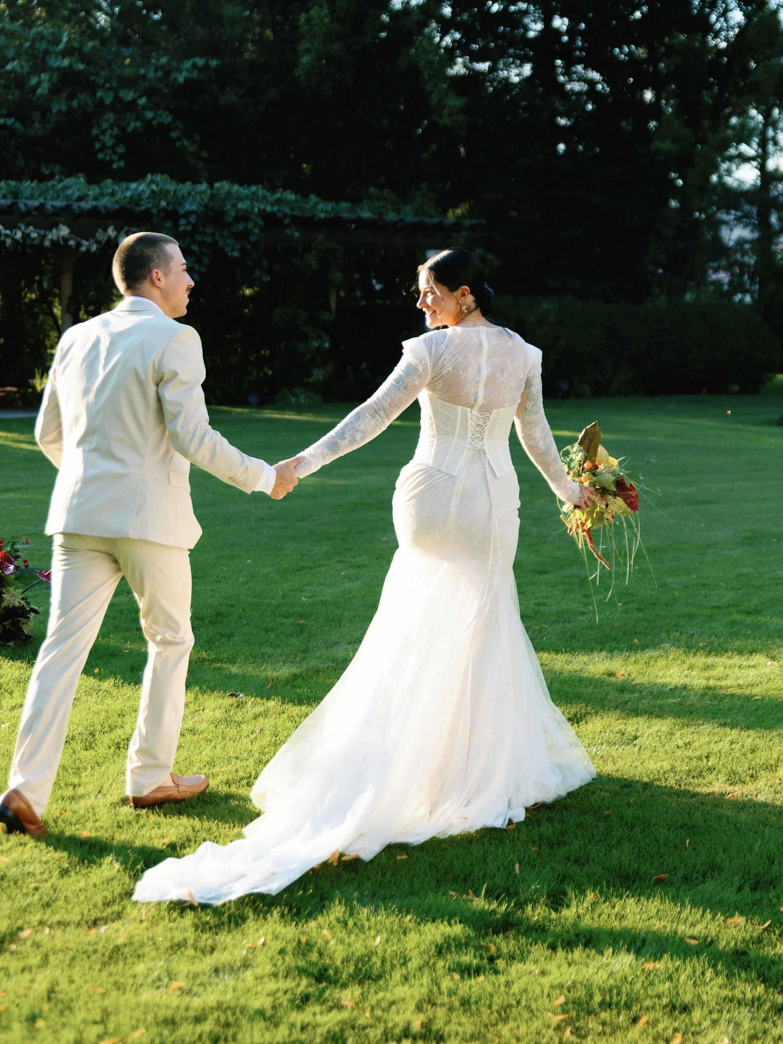 A bride and groom holding hands on a green lawn in sunlight, with trees in the background. The bride is wearing a white wedding gown and holding a bouquet, while the groom is dressed in a light-colored suit.