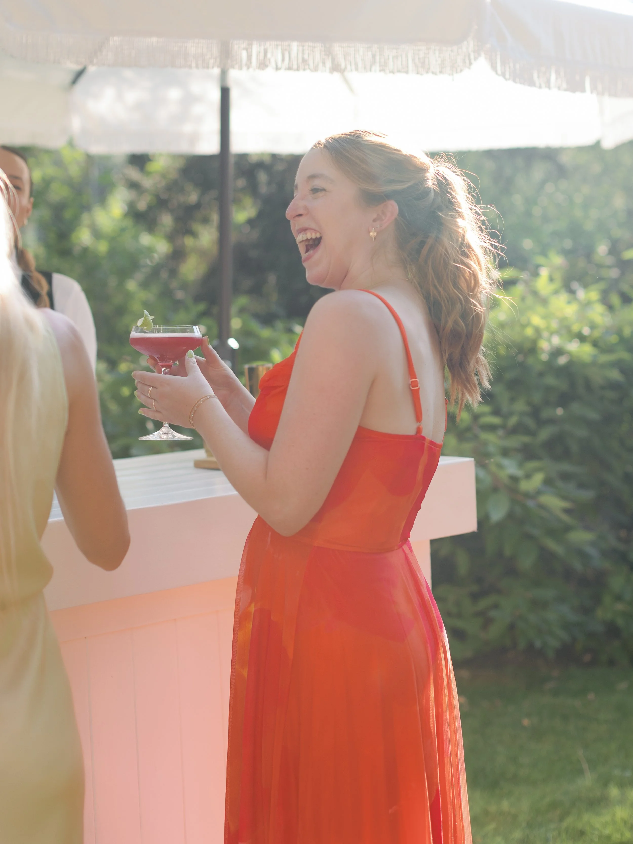 A woman in a red dress holding a cocktail and smiling at a social gathering outdoors.
