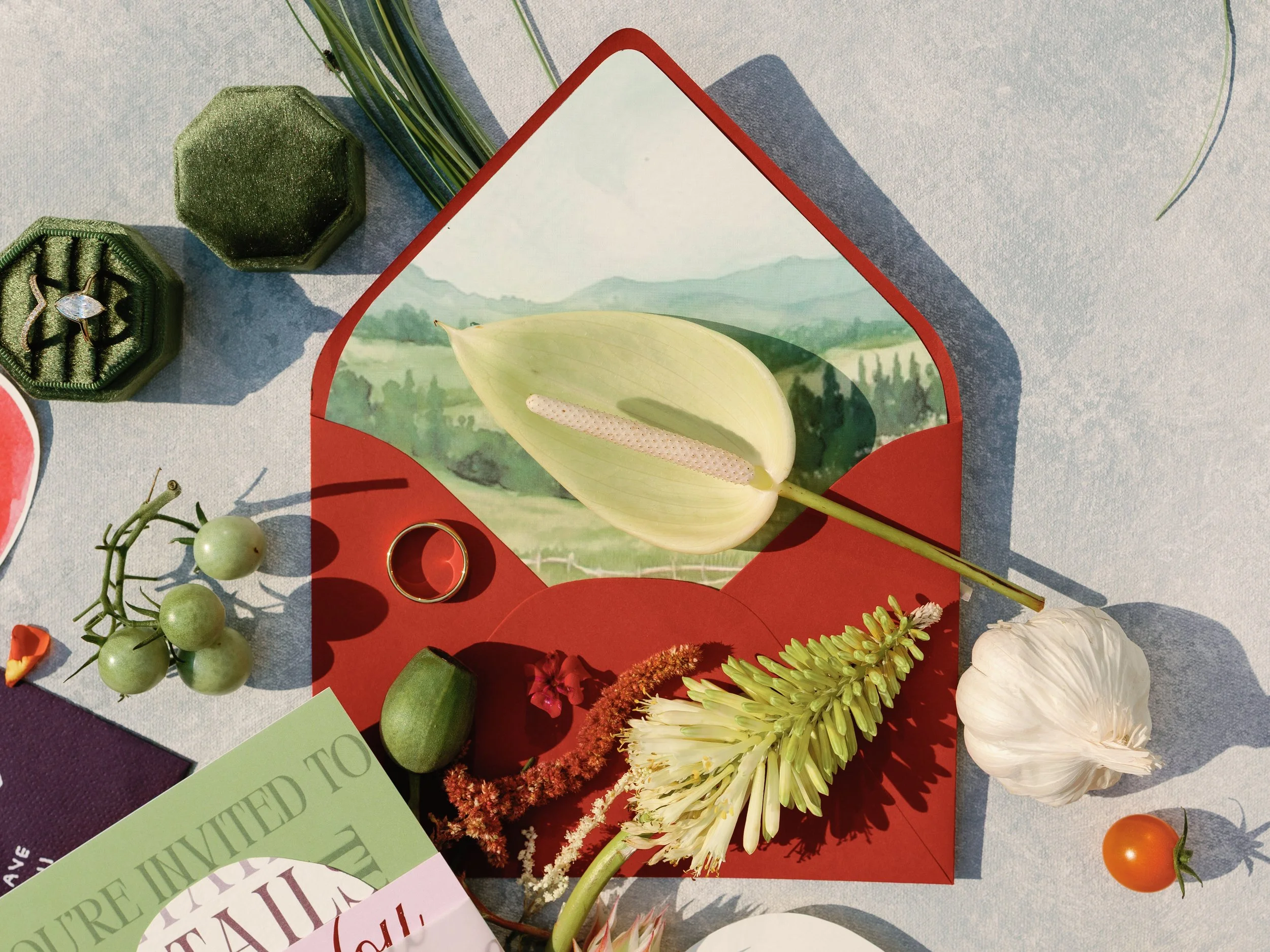 A floral arrangement with white and green flowers, a garlic bulb, and cherry tomatoes on a table with a red and green card and jewelry in green velvet boxes.