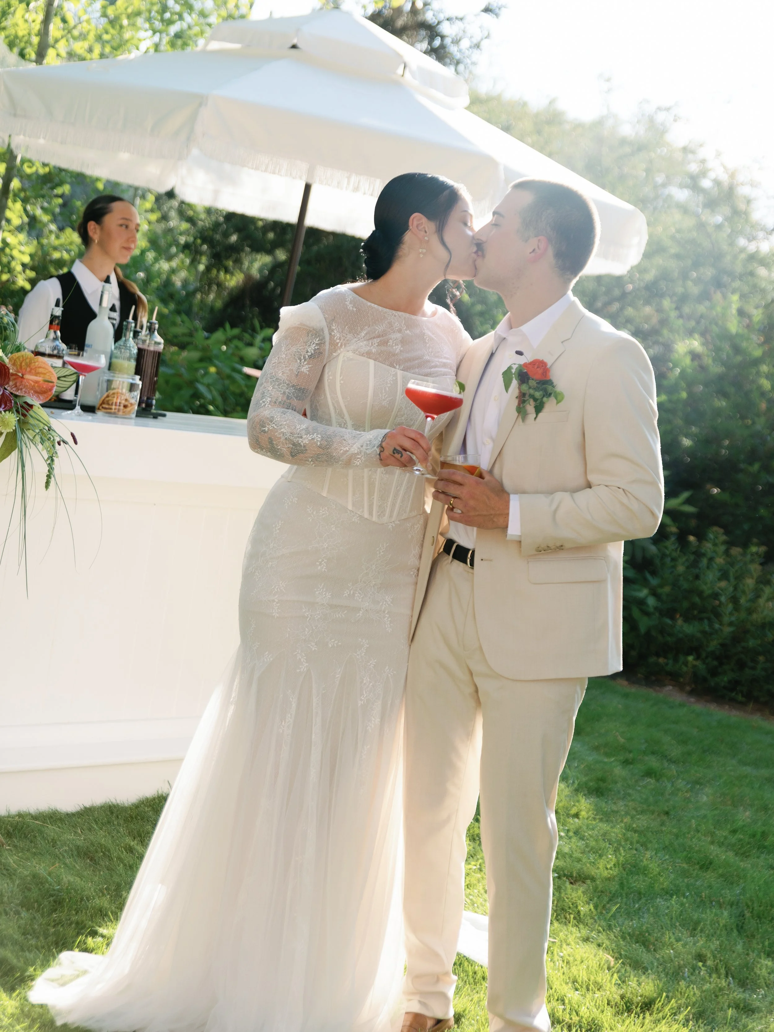 A bride and groom in wedding attire sharing a kiss outdoors, holding drinks, with a bartender and bar setup under a white umbrella in the background.