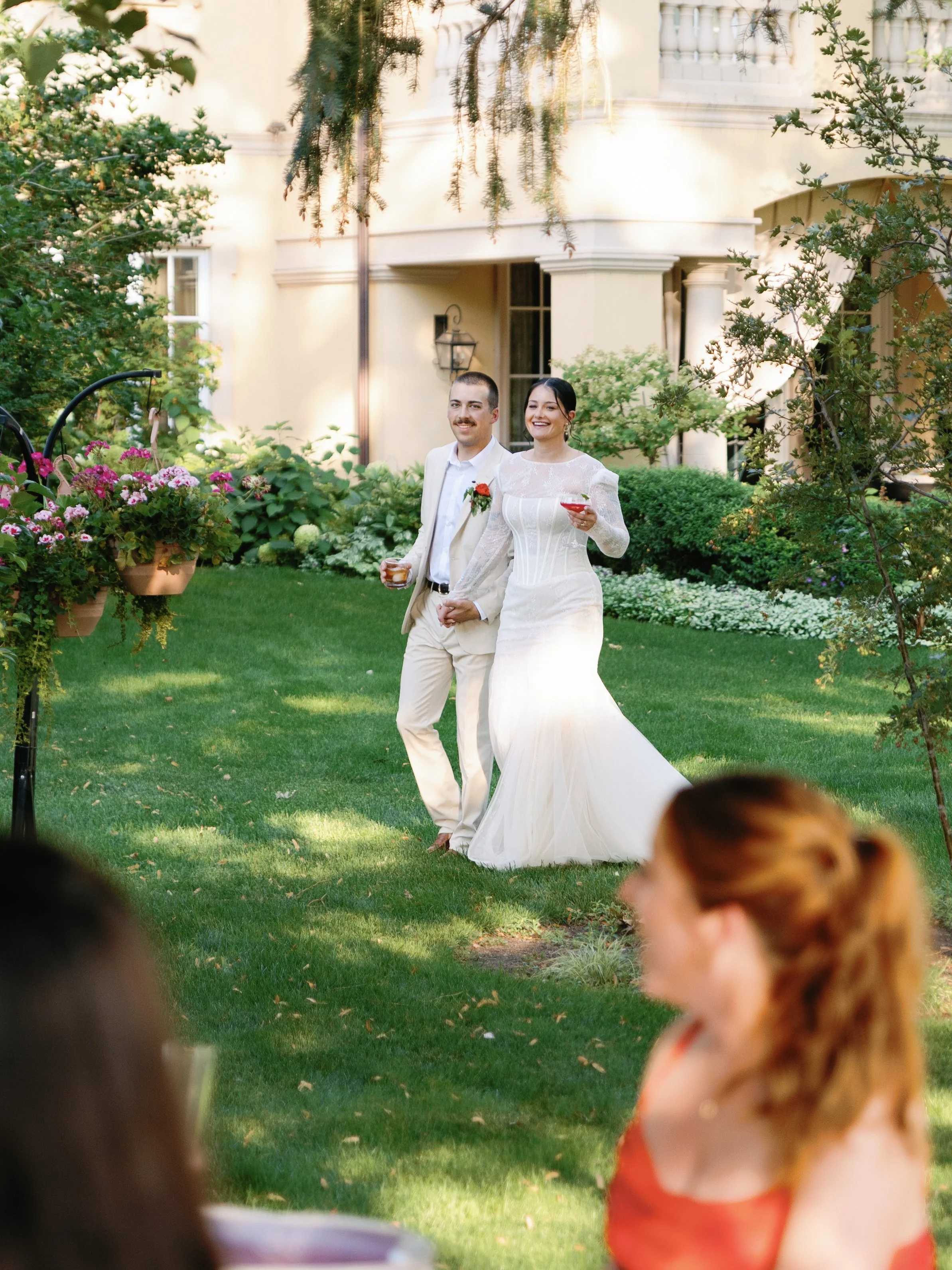 A newlywed couple walking hand in hand outdoors in a garden setting, smiling, as guests watch. The bride is wearing a white wedding gown, and the groom is in a light suit. The scene is sunny and lush with greenery.