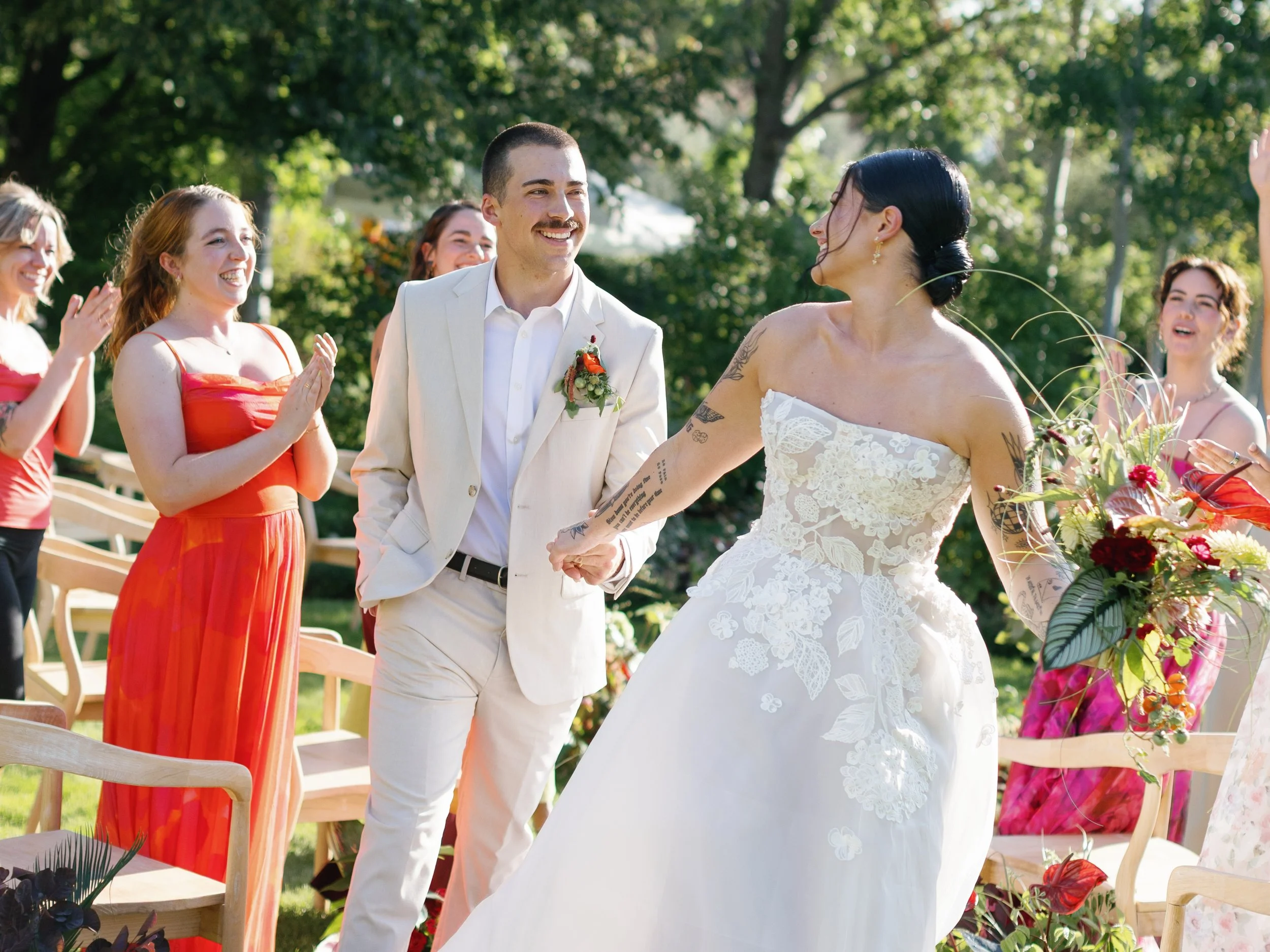 Bride and groom holding hands and smiling at each other during an outdoor wedding ceremony, with guests clapping and celebrating around them, set in a green park.