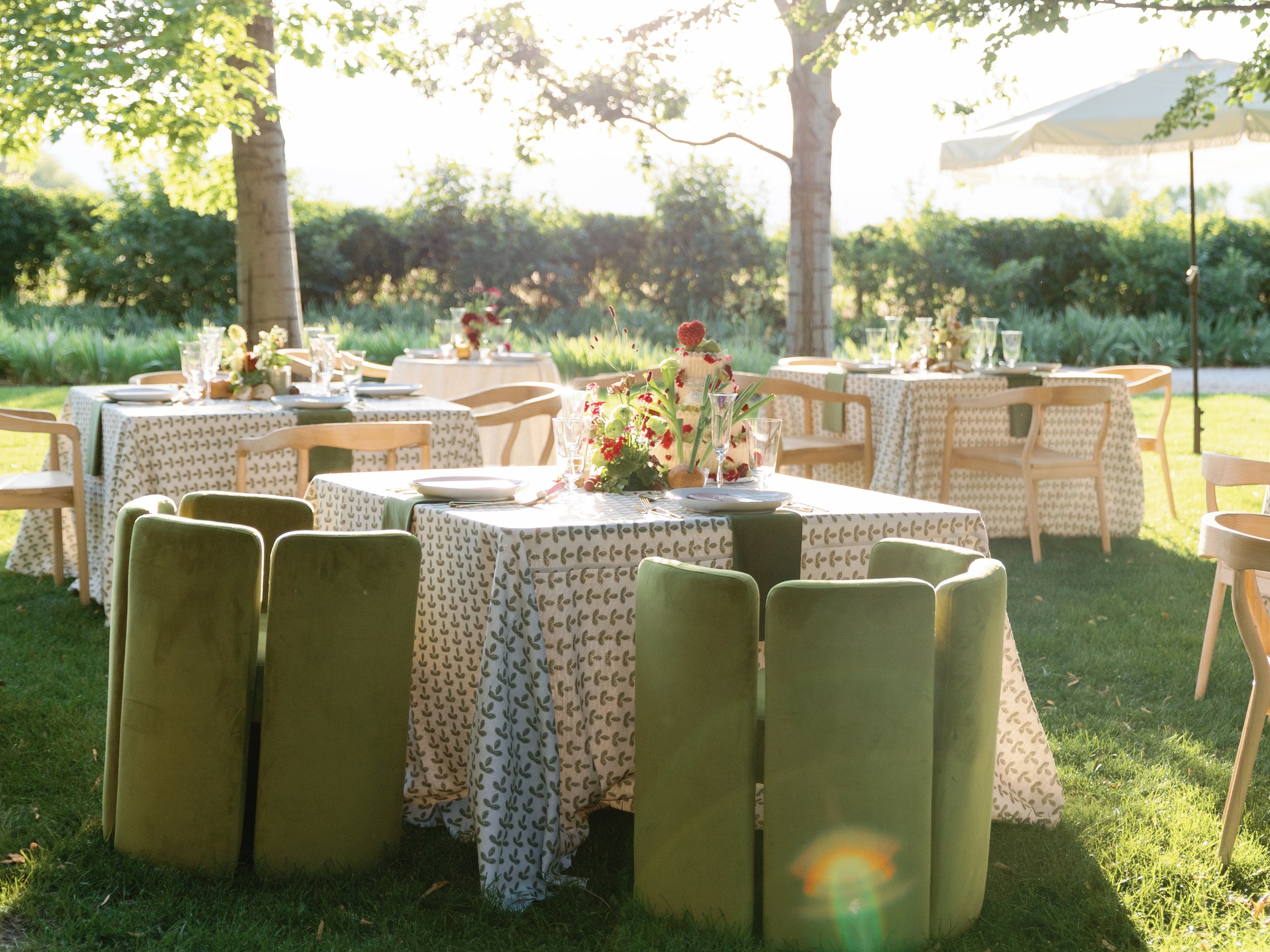 Outdoor wedding reception setup with decorated tables, chairs, and a large white umbrella shaded area on a grassy lawn with trees and greenery in the background.