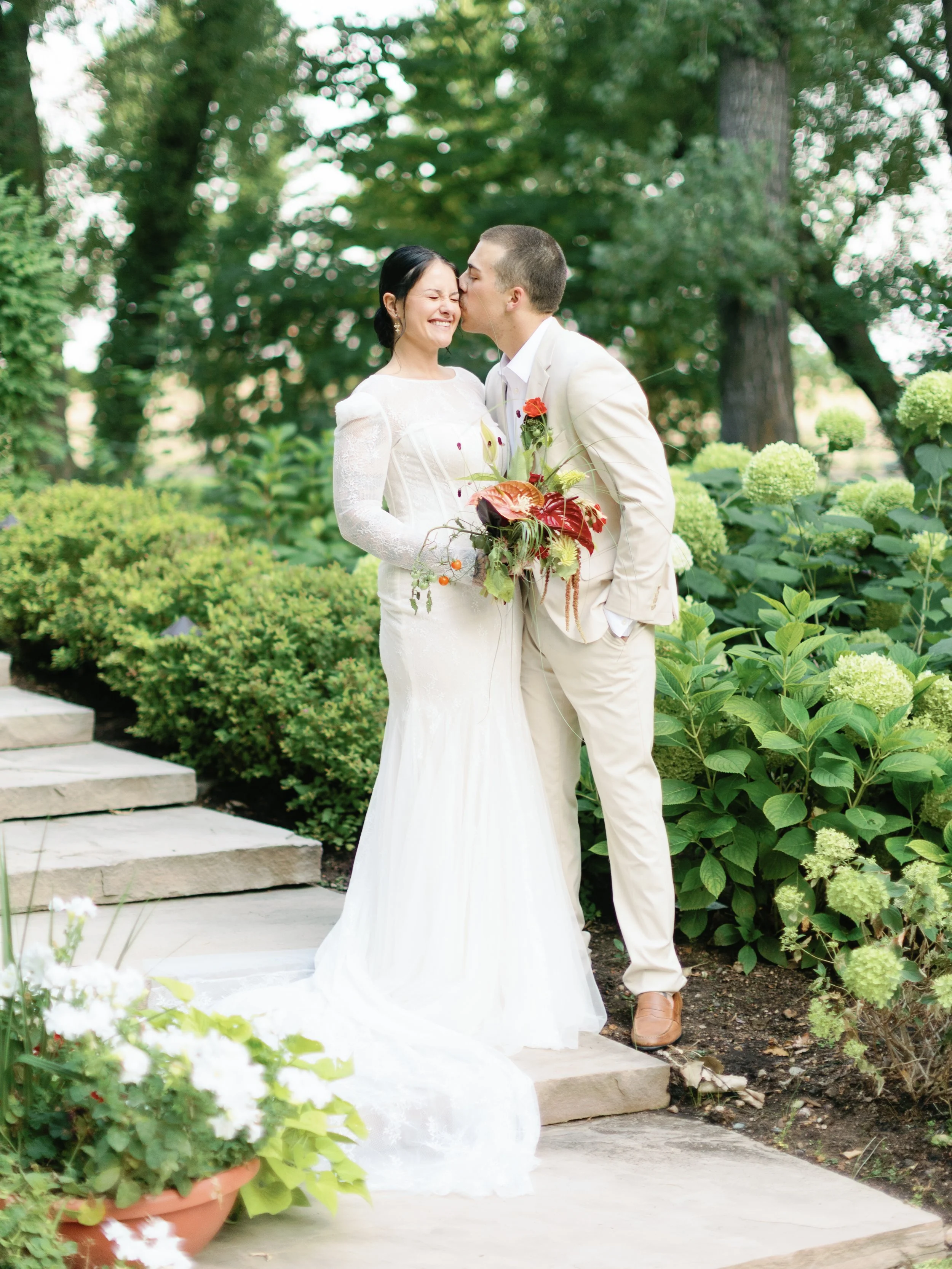 A newlywed couple sharing a moment outdoors, with greenery and white flowers around them. The bride is wearing a white wedding dress, and the groom is in a light-colored suit. The groom is kissing the bride on the cheek.