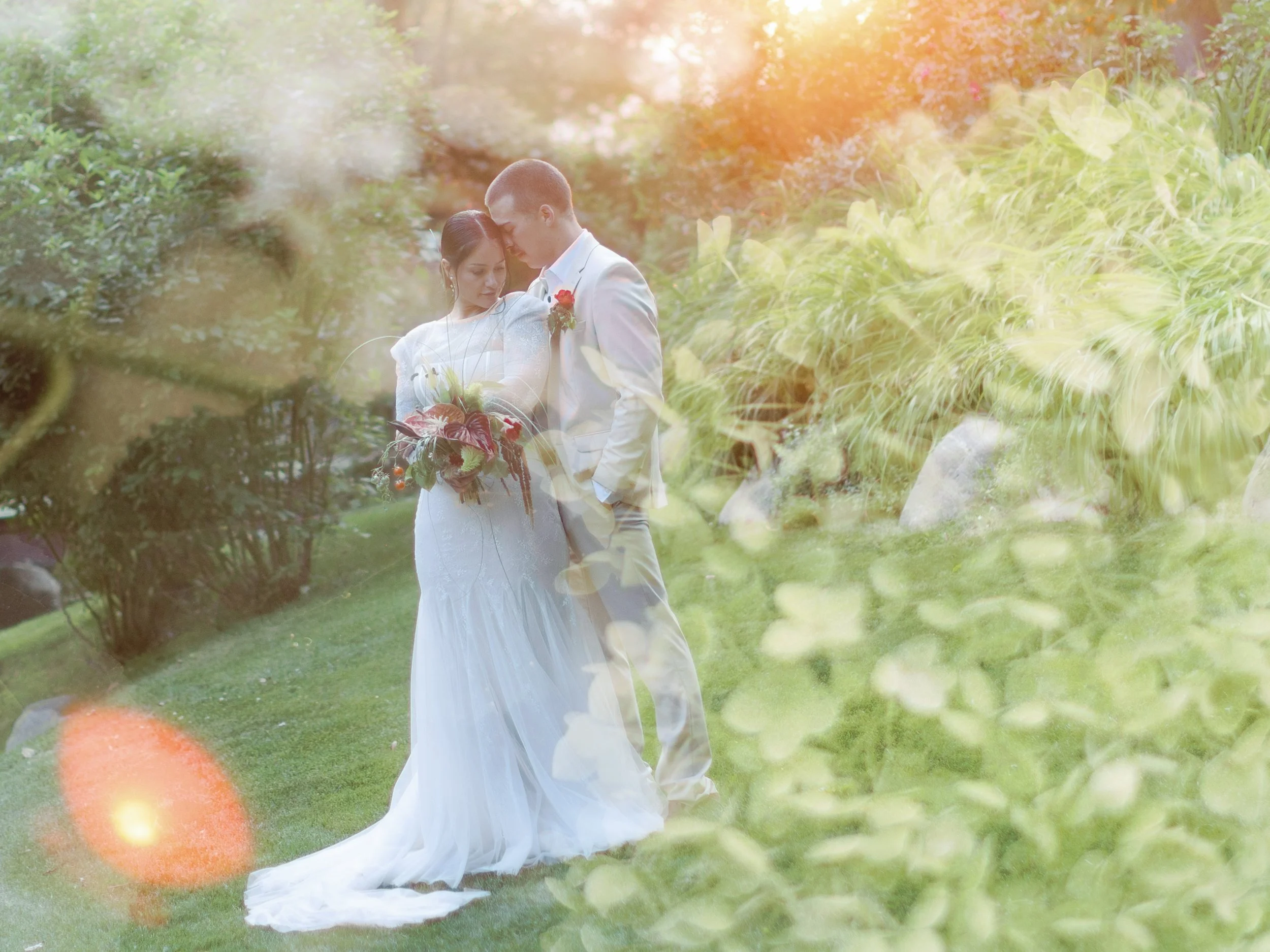 A bride and groom embracing outdoors during a wedding, with greenery and sunlight creating a soft, romantic atmosphere.