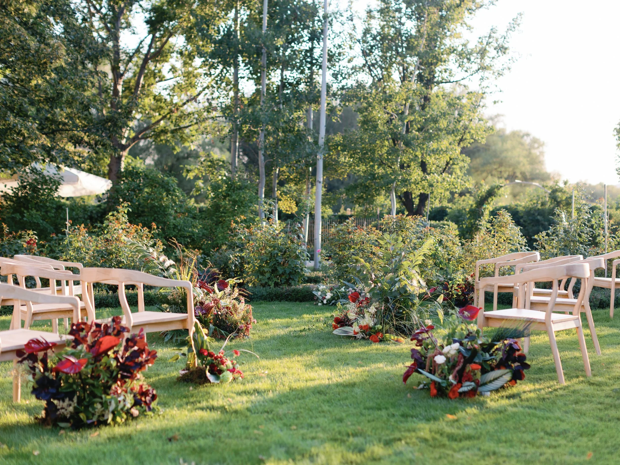 Outdoor garden setting with wooden benches arranged on green grass and colorful flower arrangements in the foreground. Tall trees and shrubs in the background, illuminated by sunlight.