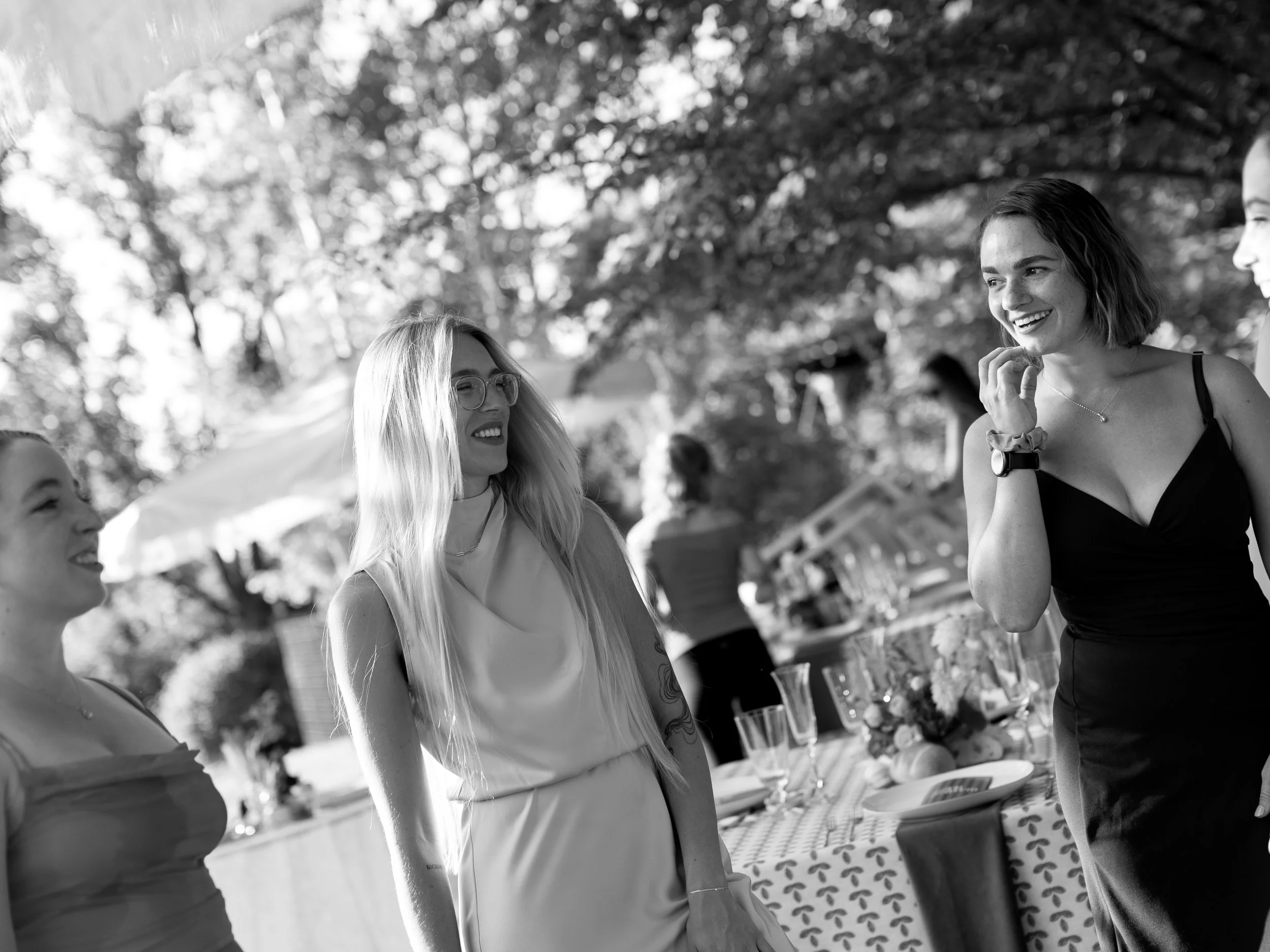 Four women talking and smiling at an outdoor event with a table and drinks in the background.