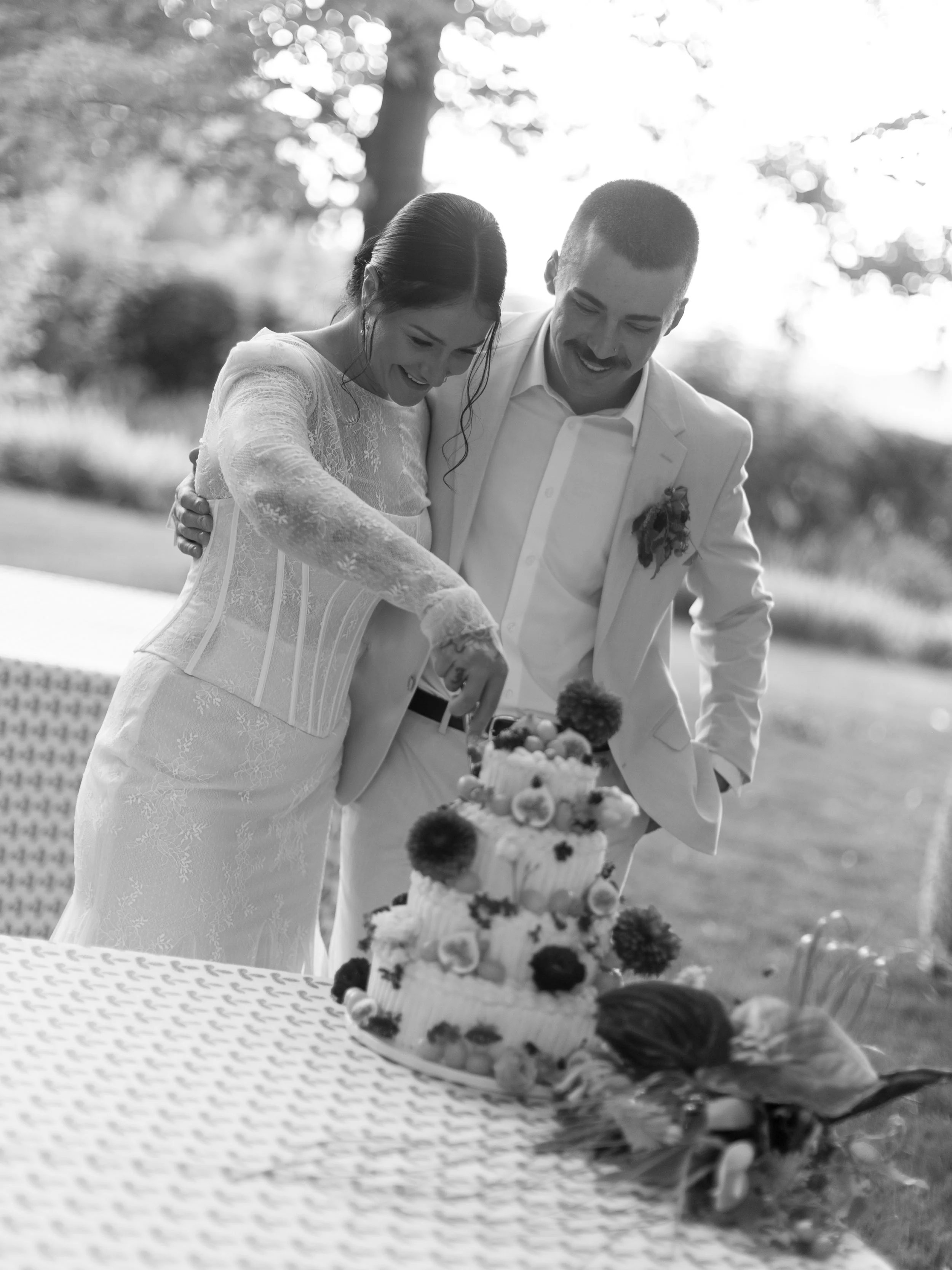 A bride and groom slicing a wedding cake outdoors during the daytime, smiling and sharing a joyful moment.
