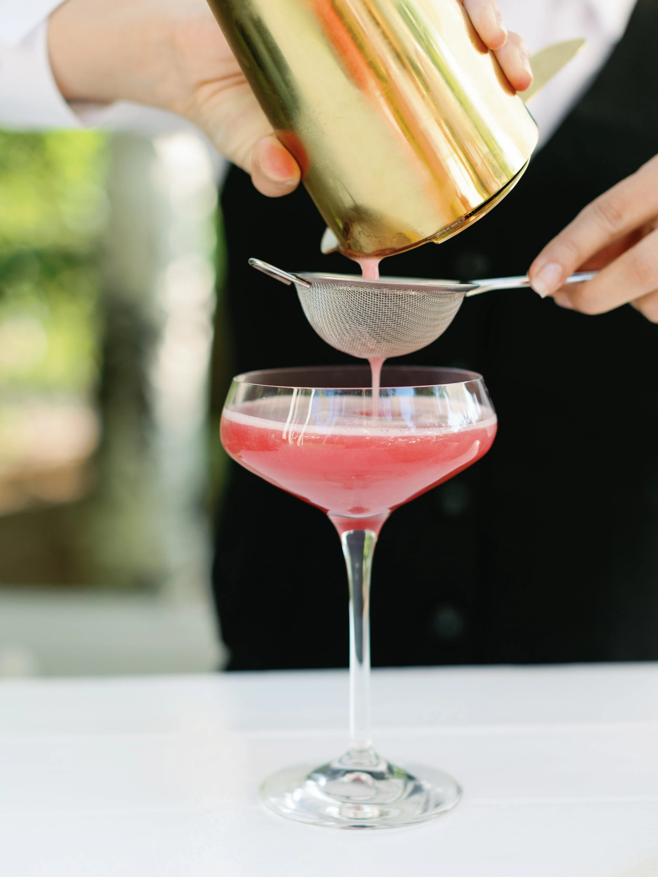 A person pouring a pink cocktail from a shaker through a strainer into a coupe glass.