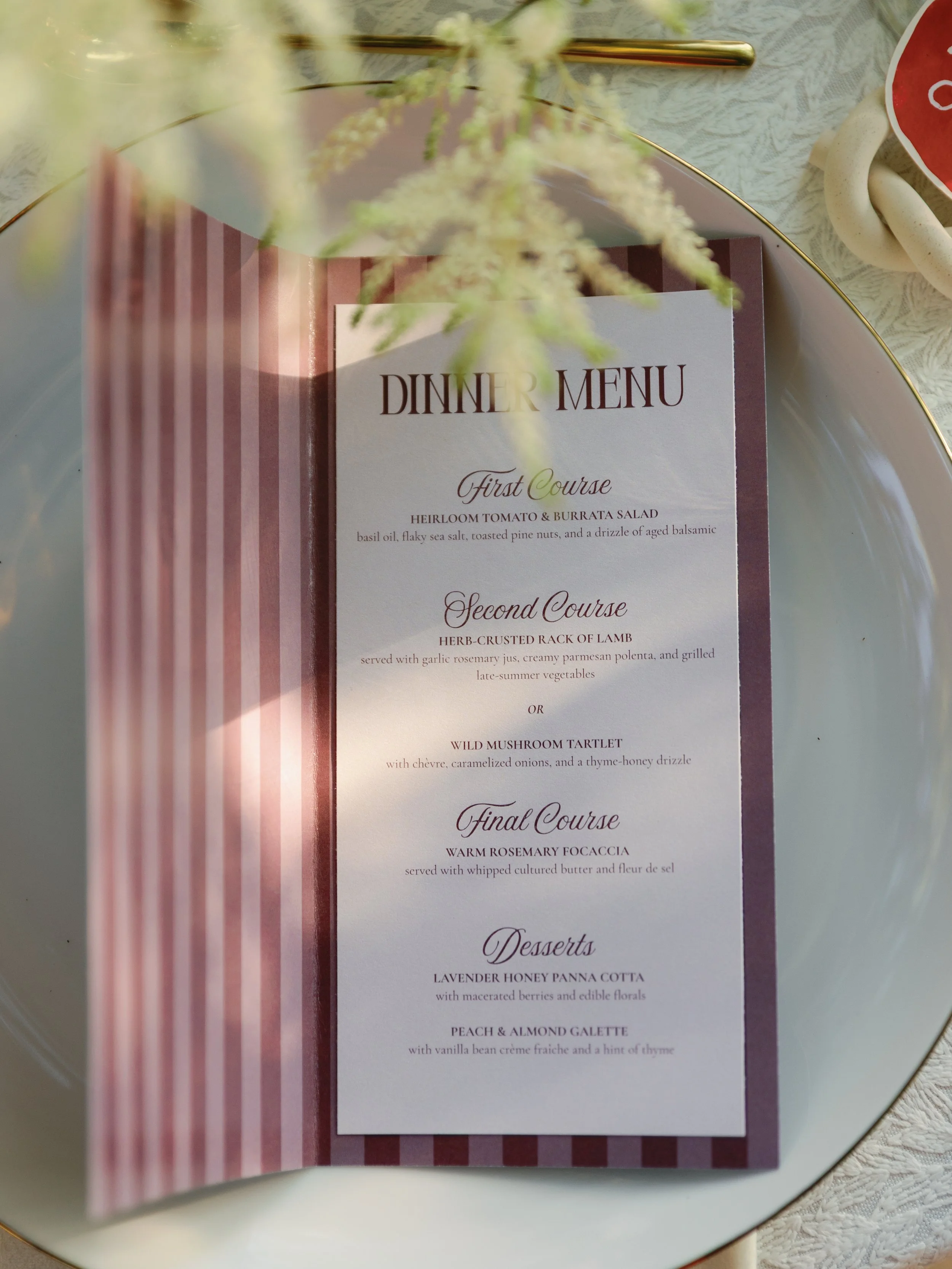A vertically folded dinner menu with a striped border, placed on a white plate. The menu lists a three-course meal with appetizers, main courses, and desserts, with elegant cursive fonts used for dish names.