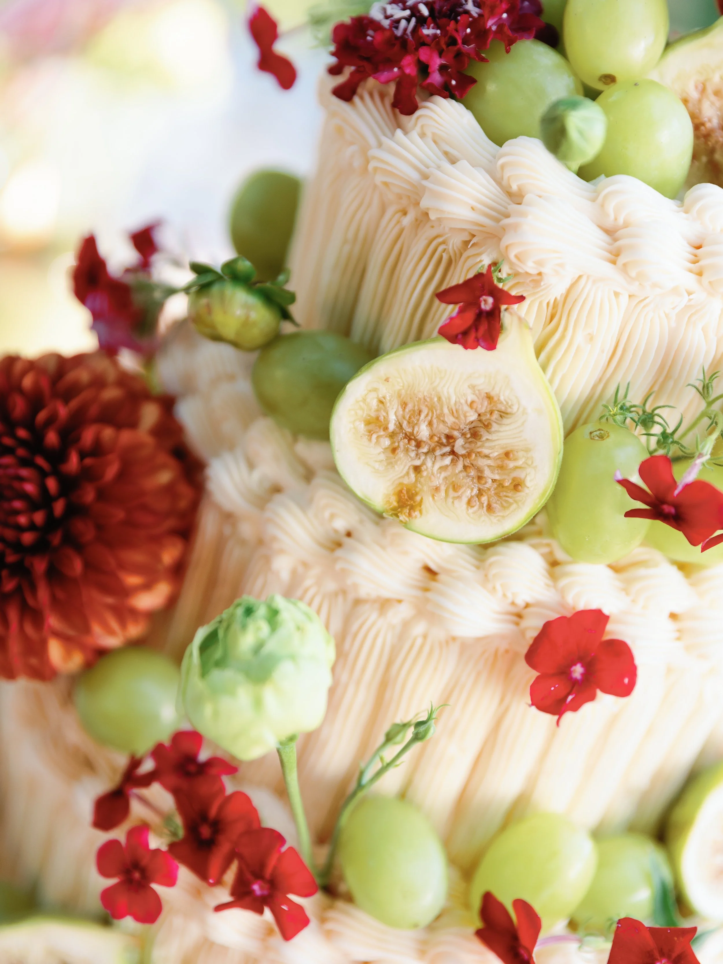 A close-up view of a cake decorated with fresh fruits including figs, green grapes, and red flowers with cream-colored icing.