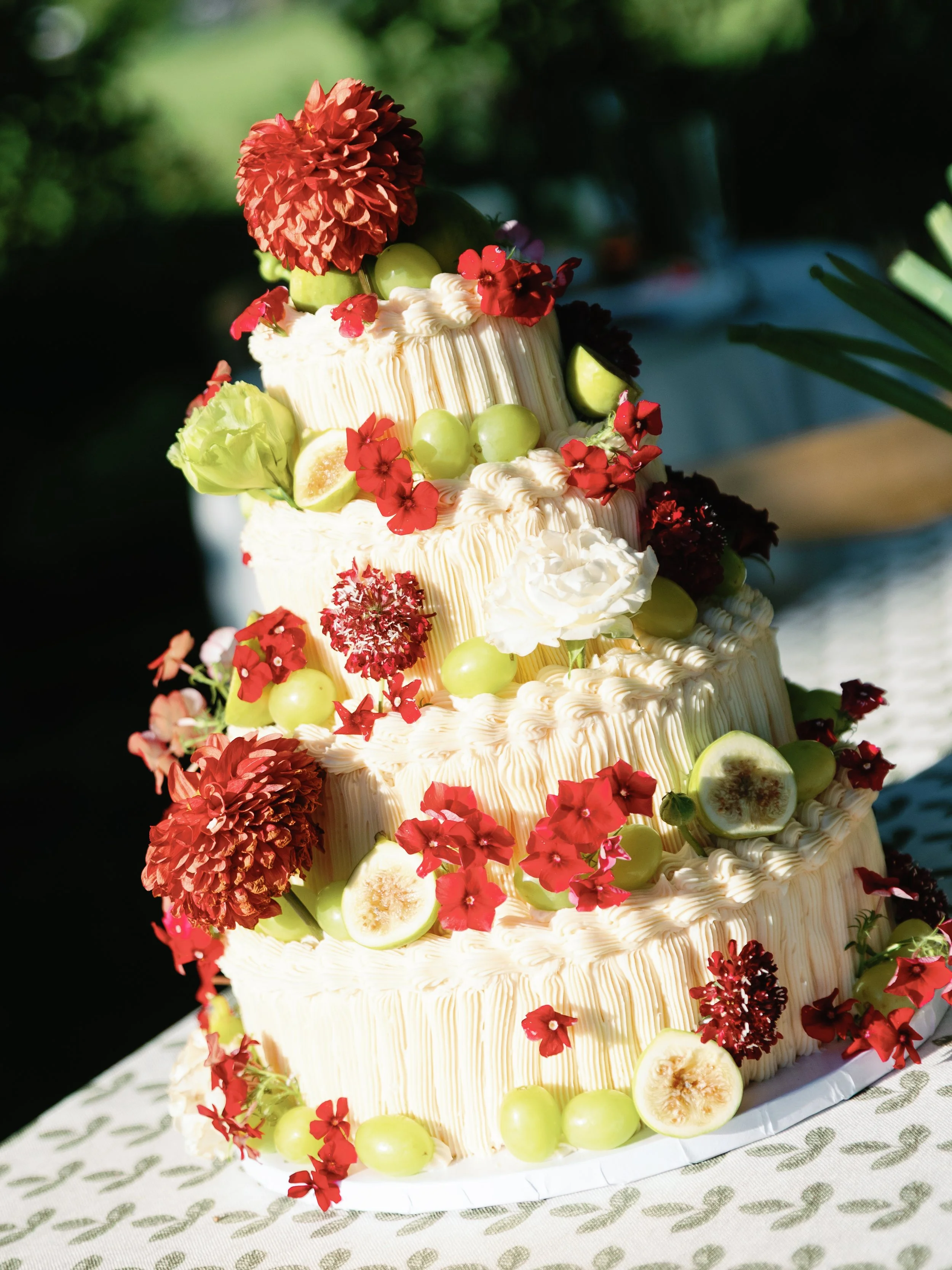 Three-tiered wedding cake decorated with white frosting, red and green flowers, green grapes, and sliced figs, set outdoors on a patterned tablecloth.