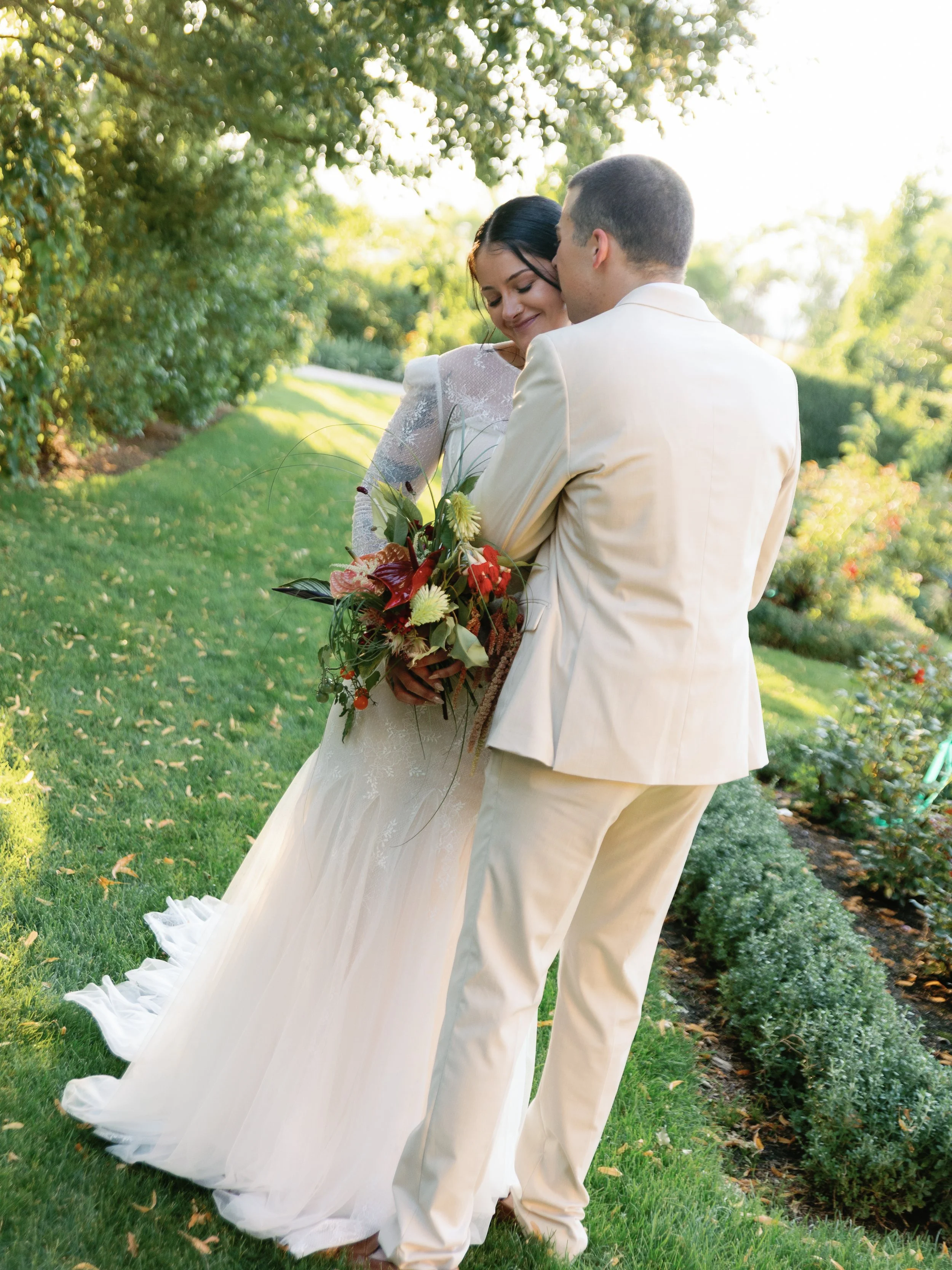 A bride and groom stand close together outdoors, surrounded by greenery, with the bride holding a large bouquet of flowers.