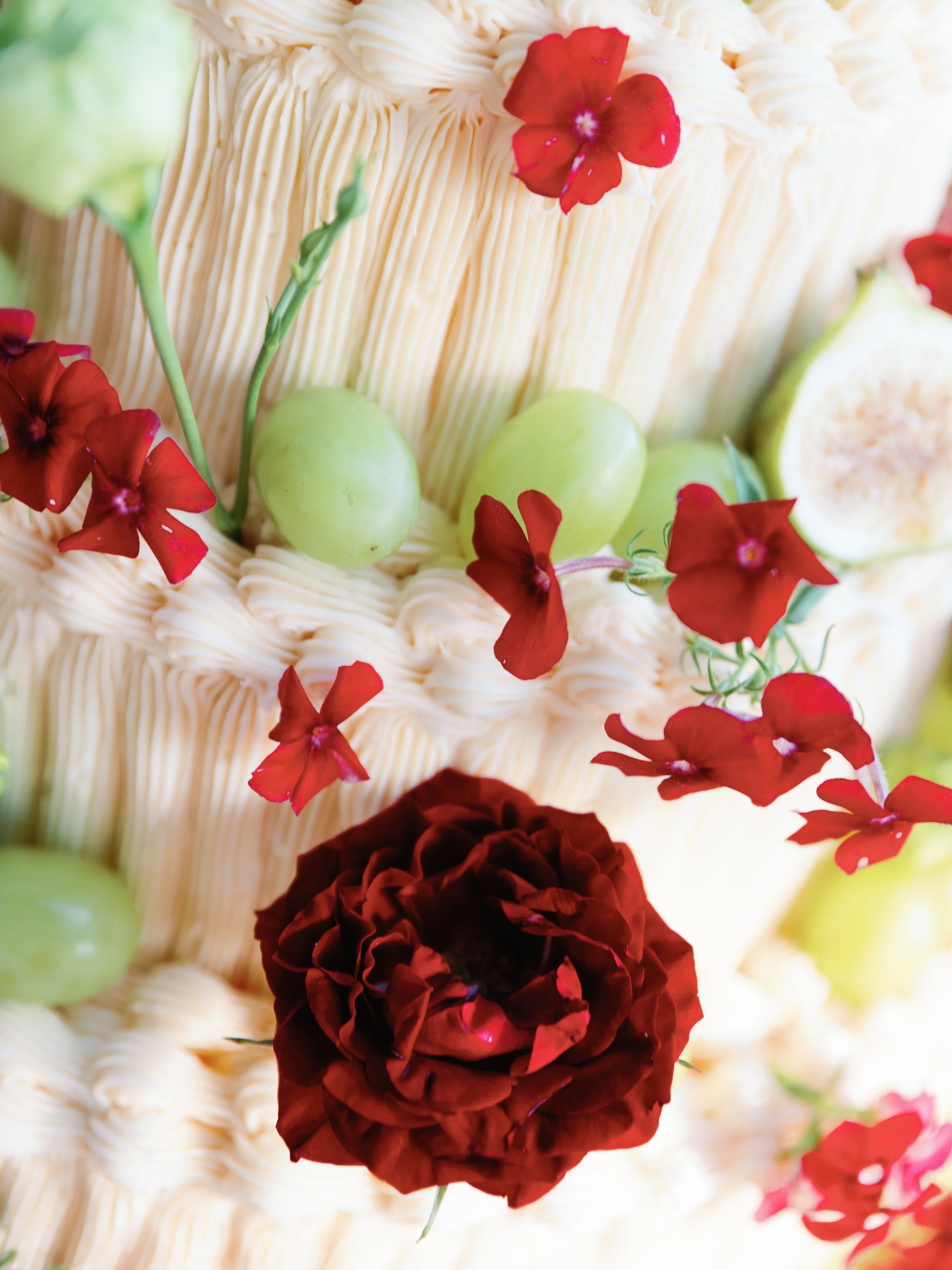 Close-up of a cake decorated with red and pink flowers, green grapes, and a sliced fig, with a textured cream-colored frosting.